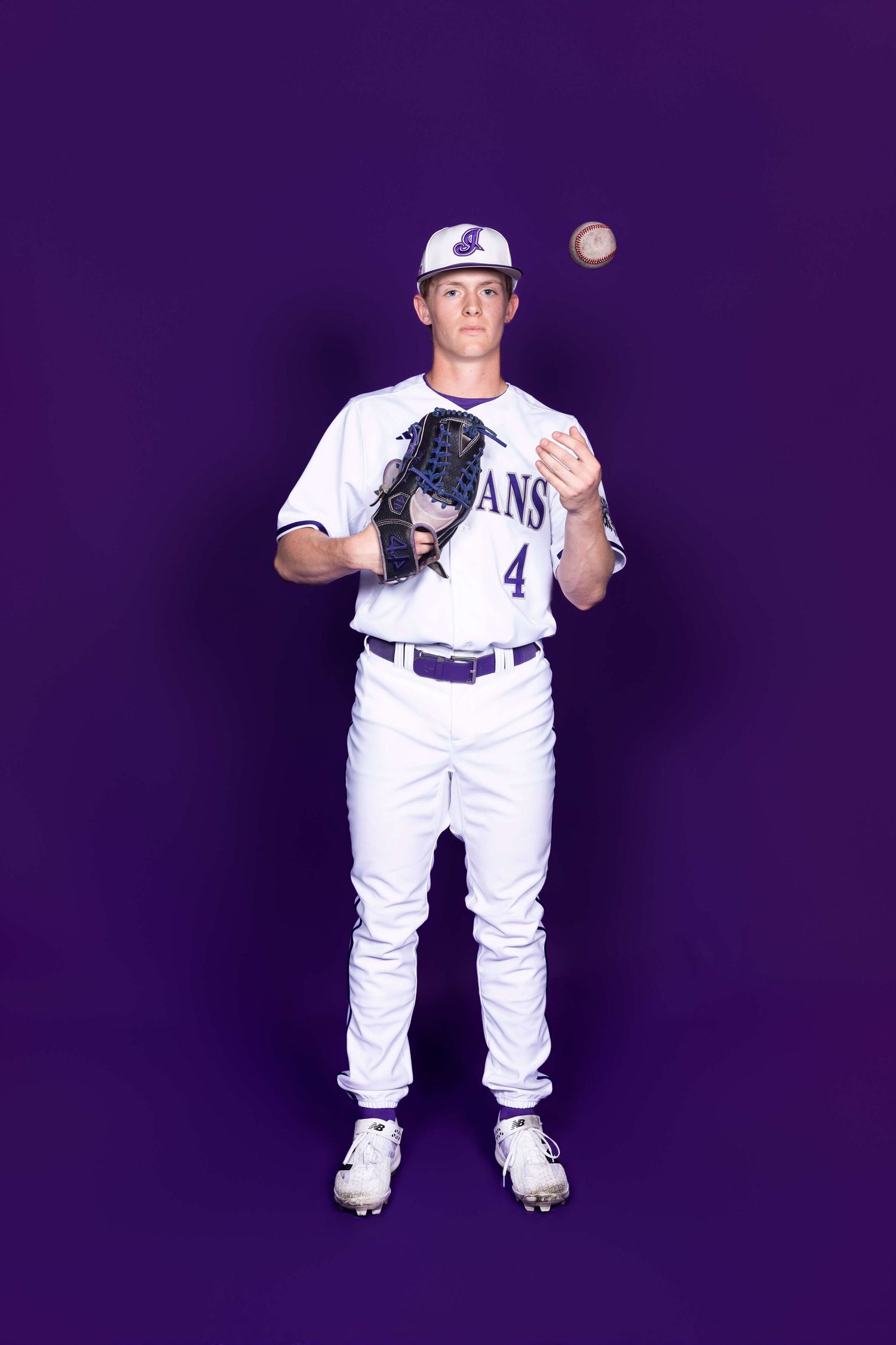 Baseball player in white uniform poses against purple background during team photo session.