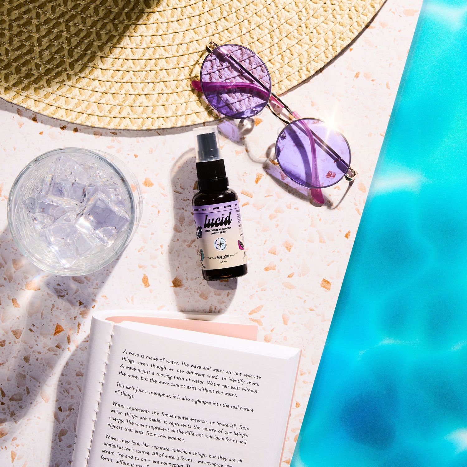 Lucid spray bottle next to purple sunglasses and book by pool with straw hat.