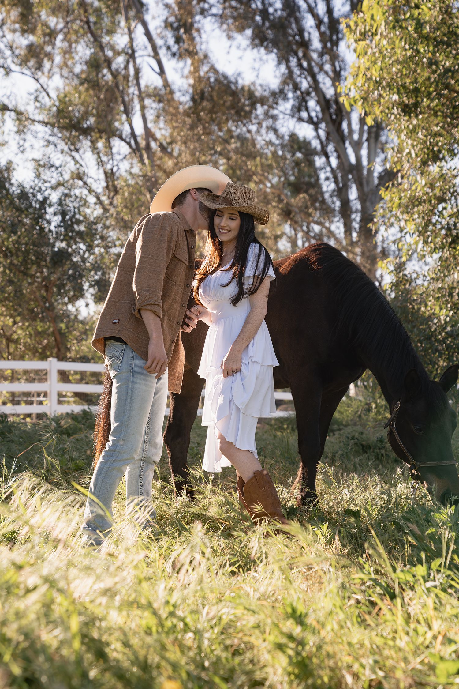 A couple and their horse stand together in a sunlit field with white fencing and trees in the background.