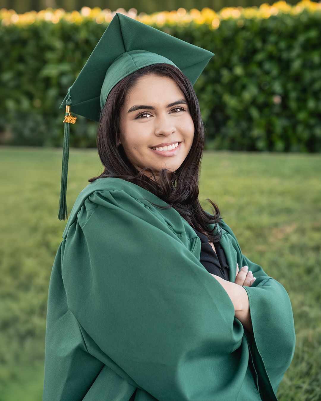 A graduate in emerald green cap and gown poses outdoors against a bokeh background.