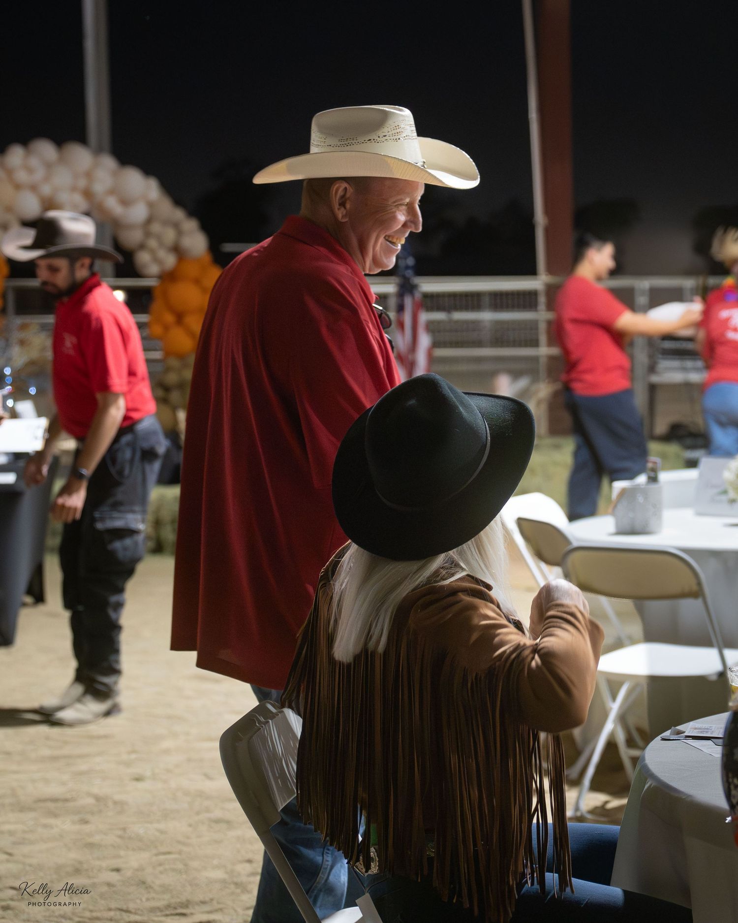 People in western attire sit at tables during an indoor event in a dimly lit venue.
