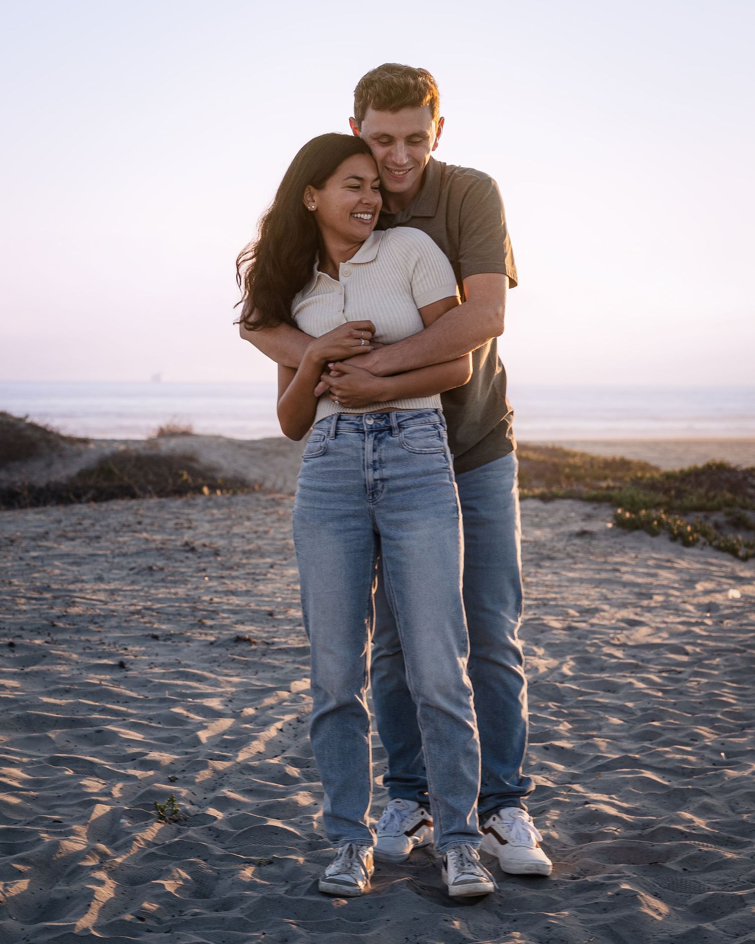 Two people stand close together on a beach wearing jeans and casual attire.