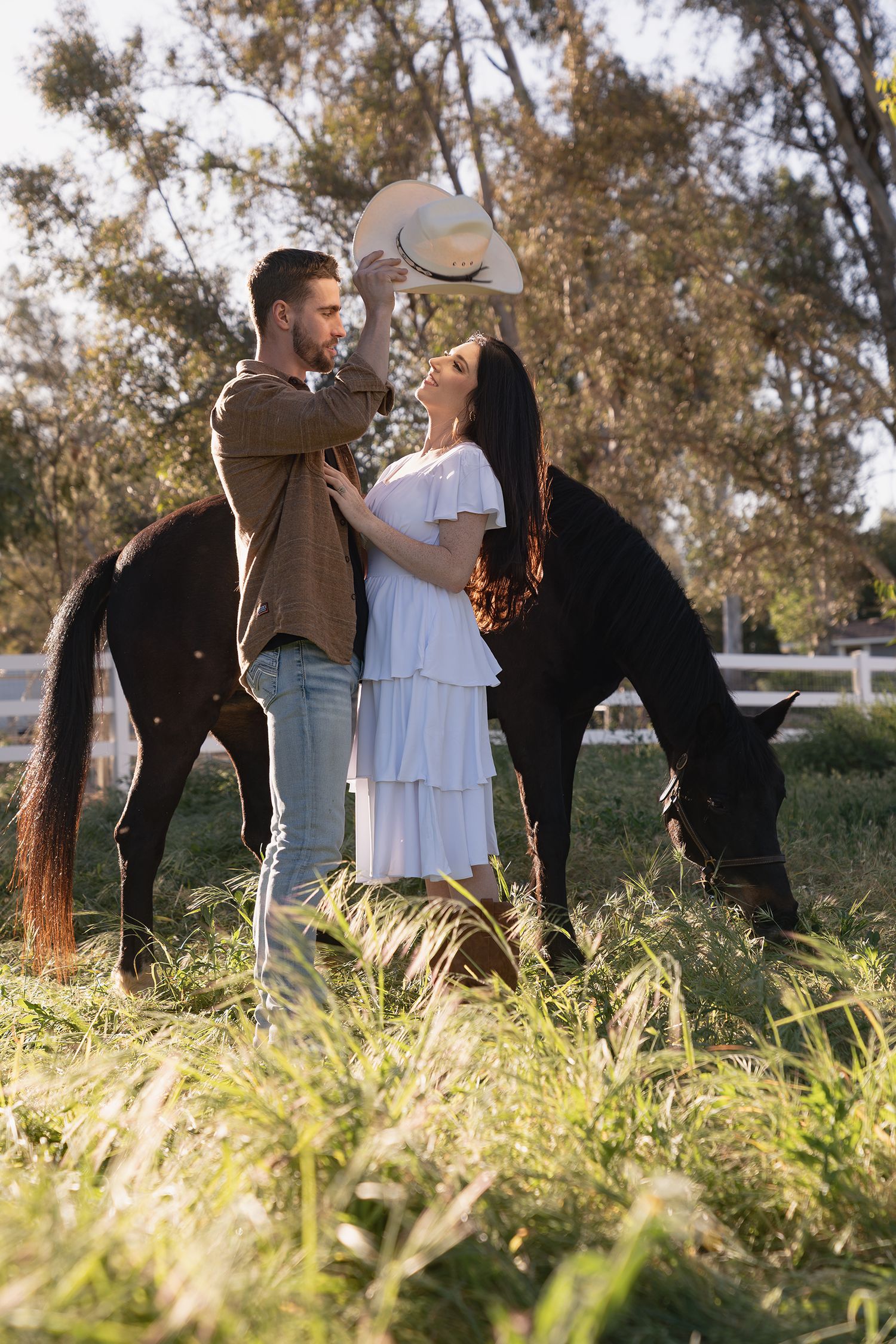 A couple in a white dress and brown suit dance together in a sunlit field of tall grass during golden hour.