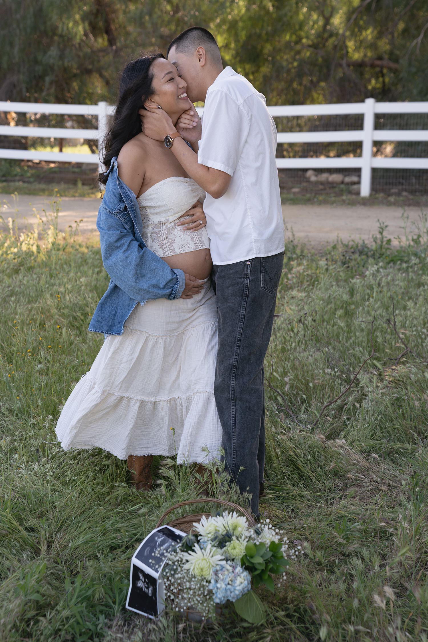 A couple embraces in a romantic outdoor setting with white fences and tall grass at sunset.