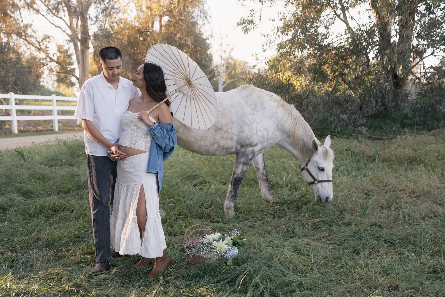 A couple in white outfits poses together during a romantic outdoor photoshoot with a white horse at sunset.
