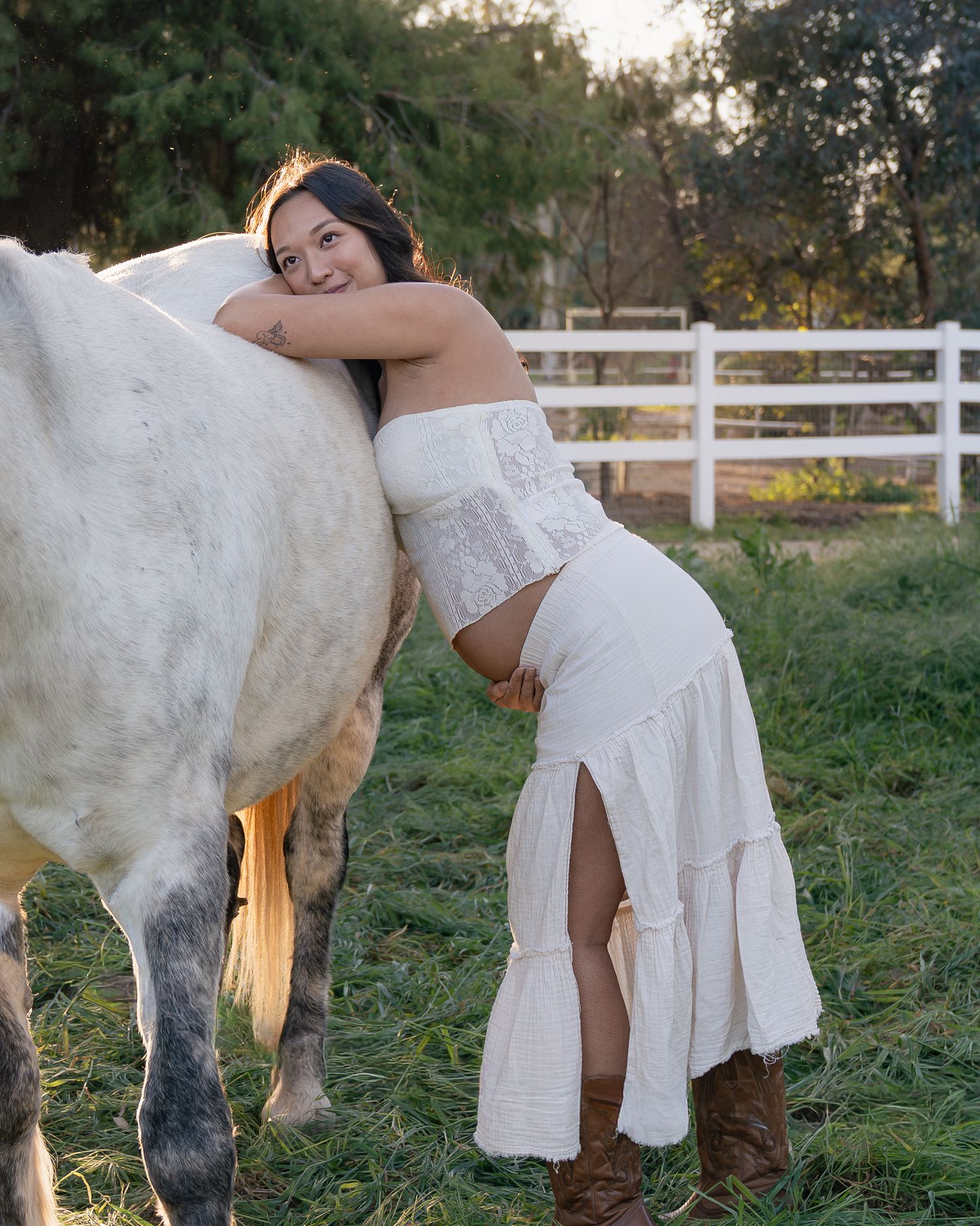 Woman in white dress hugging a white horse on a green farm with a white fence in the background.