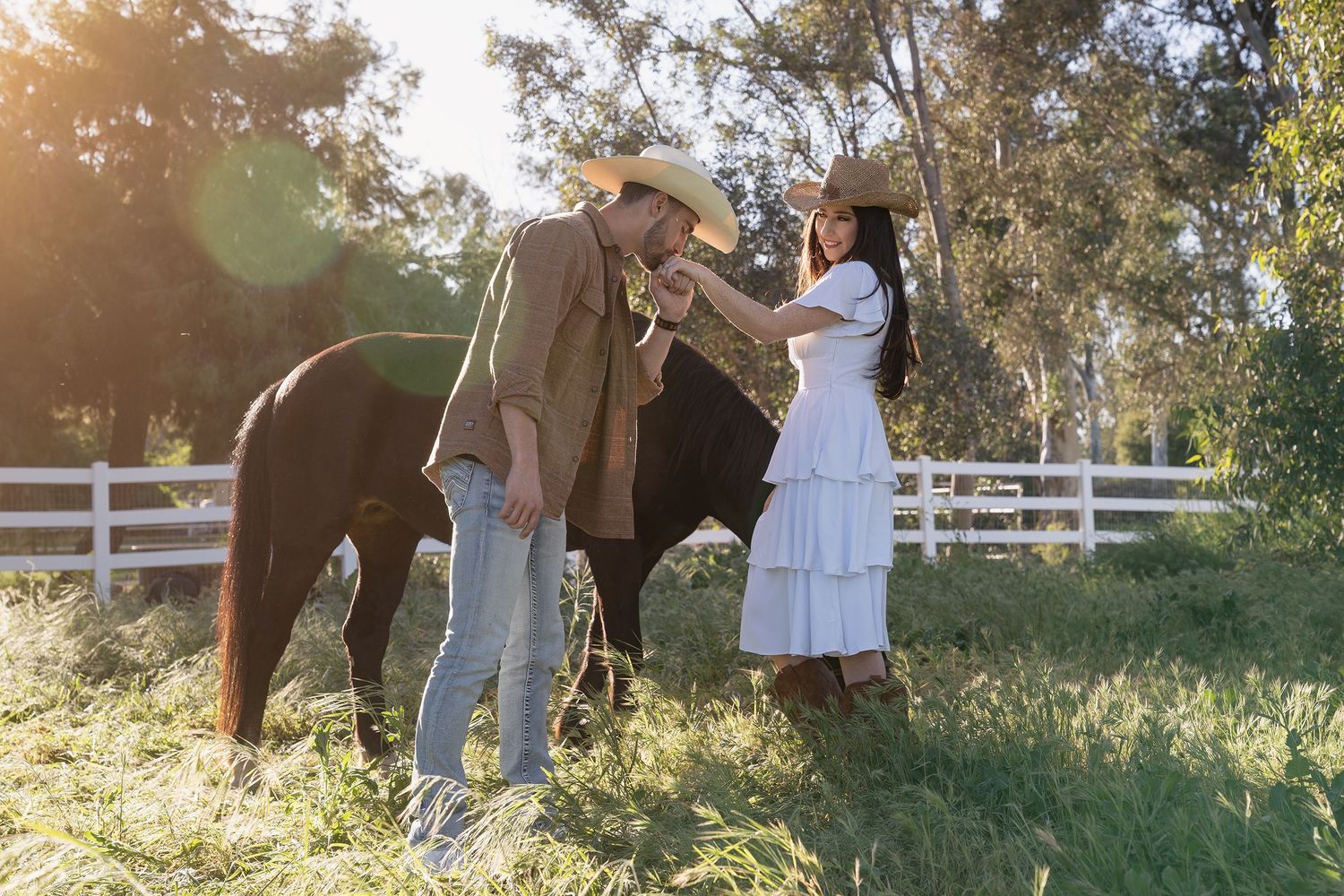 A group of people interact with brown horses in a sunlit grassy field surrounded by white fencing.