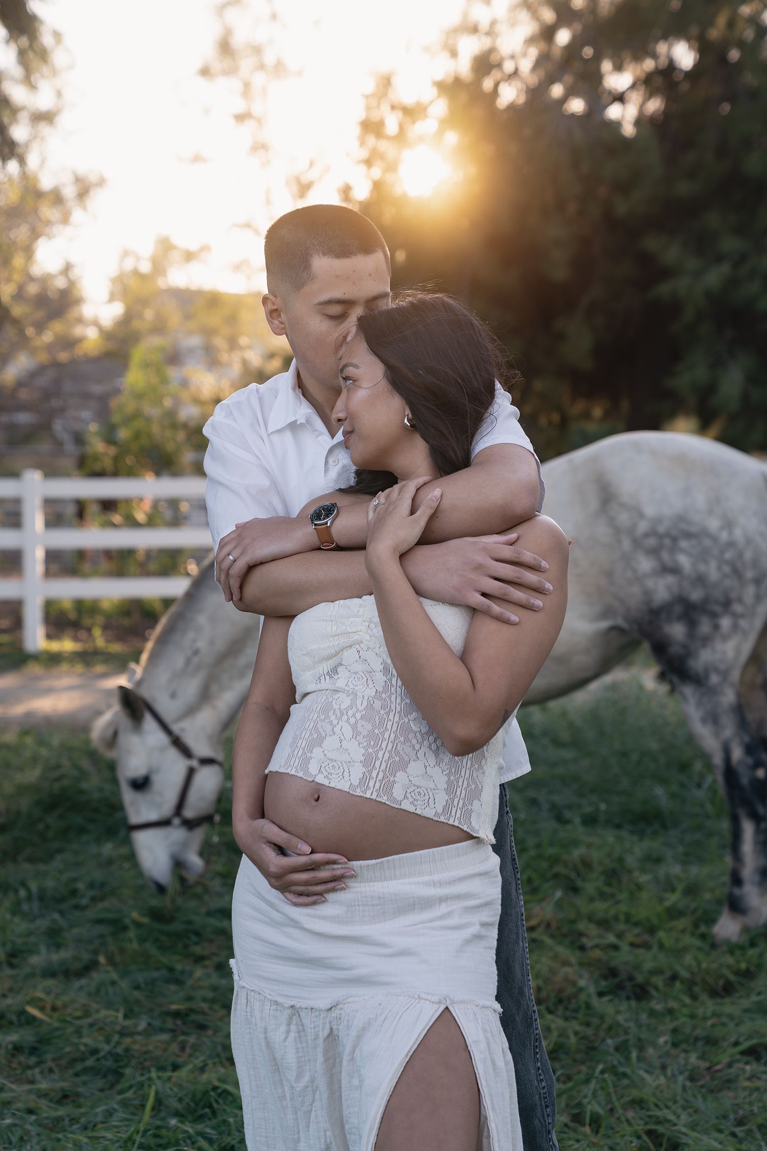 Romantic couple embraces in a sunlit field at golden hour with a white fence in the background.