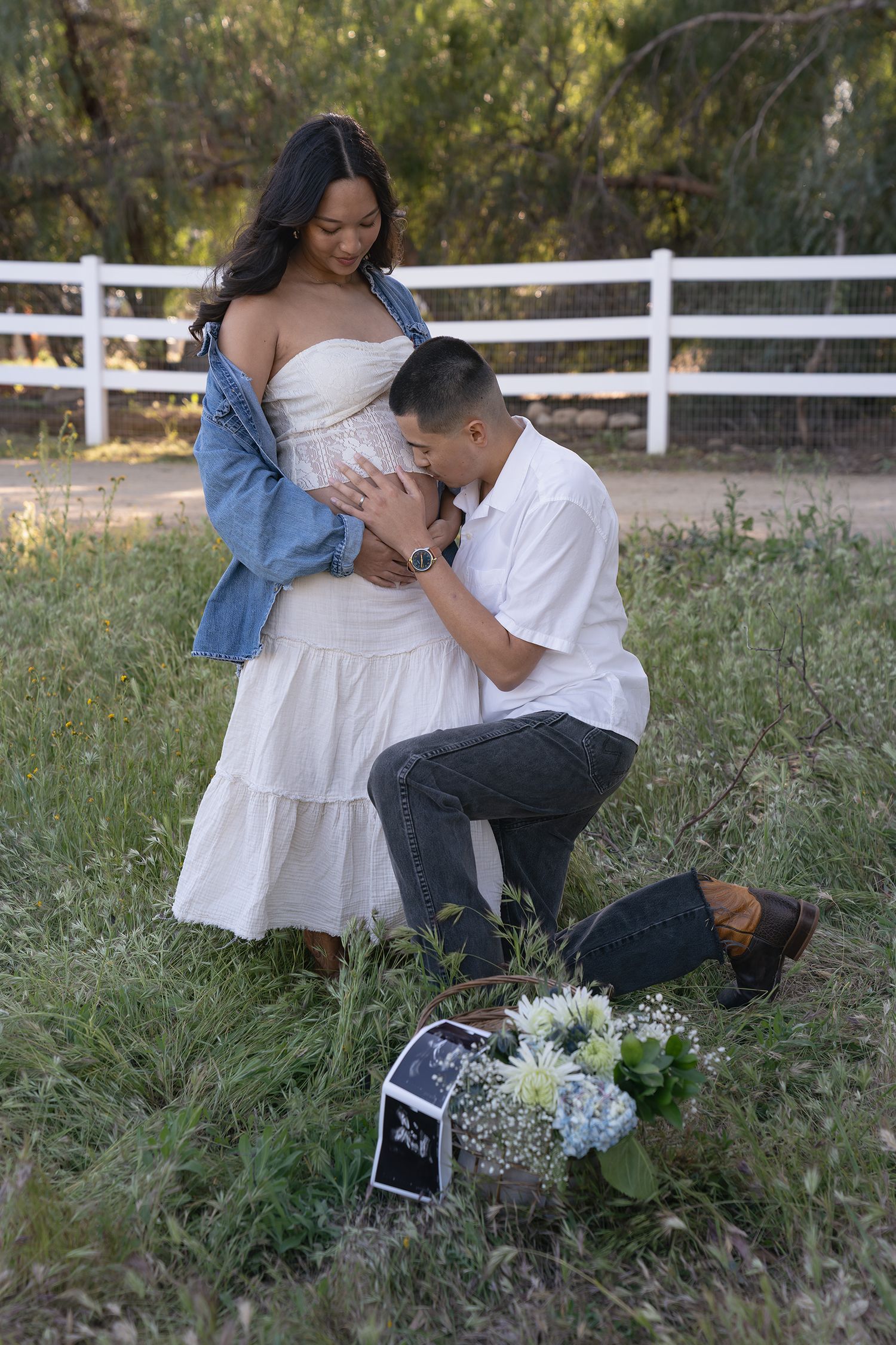A couple shares an intimate moment in a field with white fencing and a sonogram photo visible beside them.