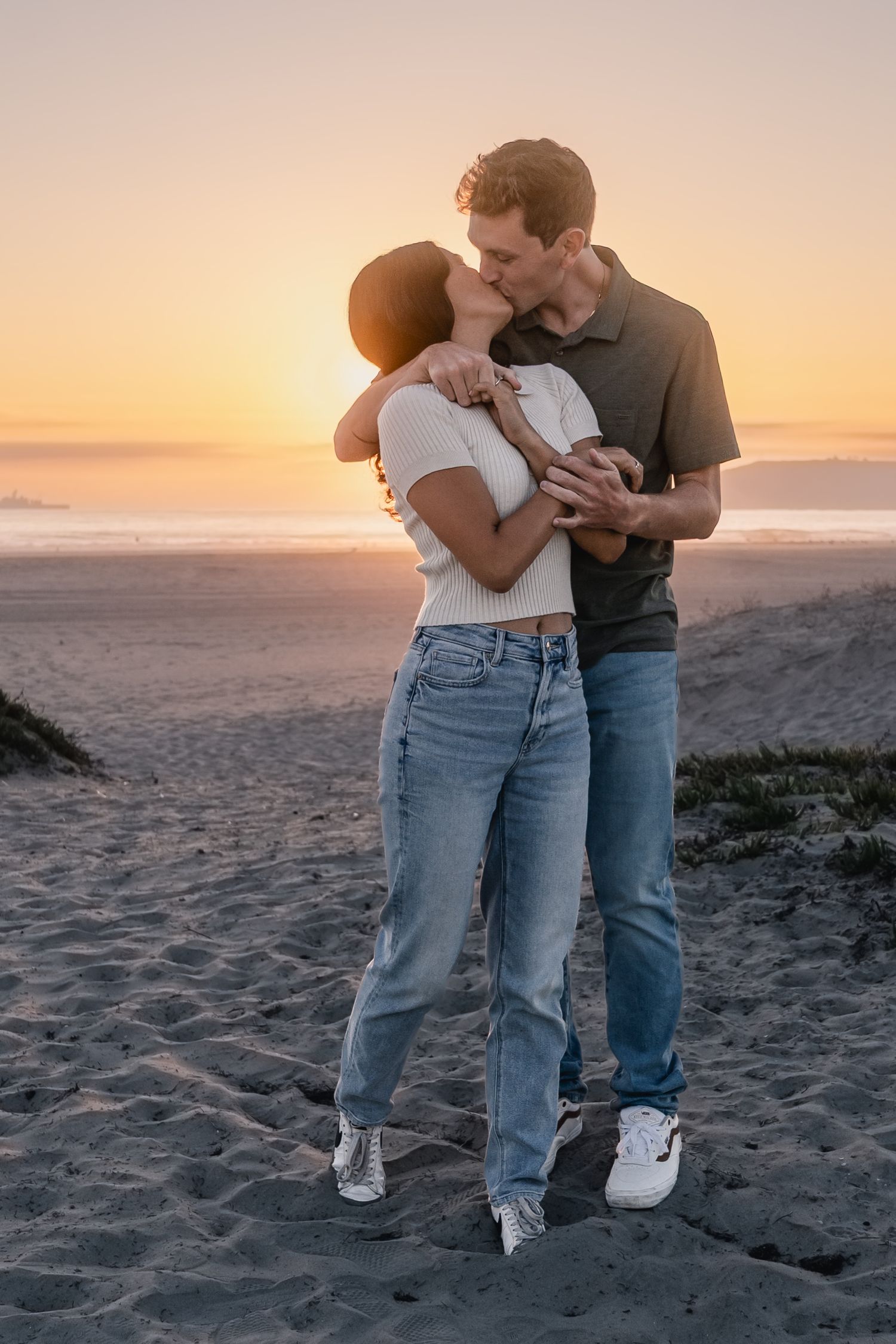 A couple shares an intimate moment on the beach at sunset wearing jeans and casual attire.