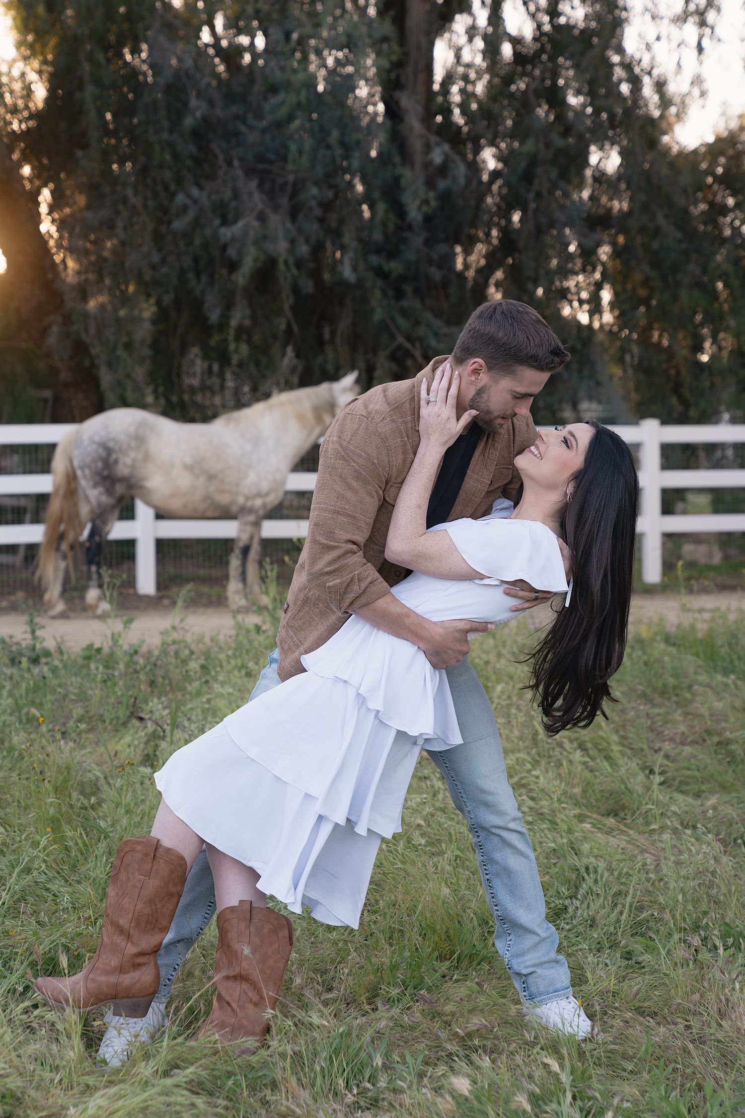 A romantic couple embraces in a field with white fence and trees in the background during golden hour.