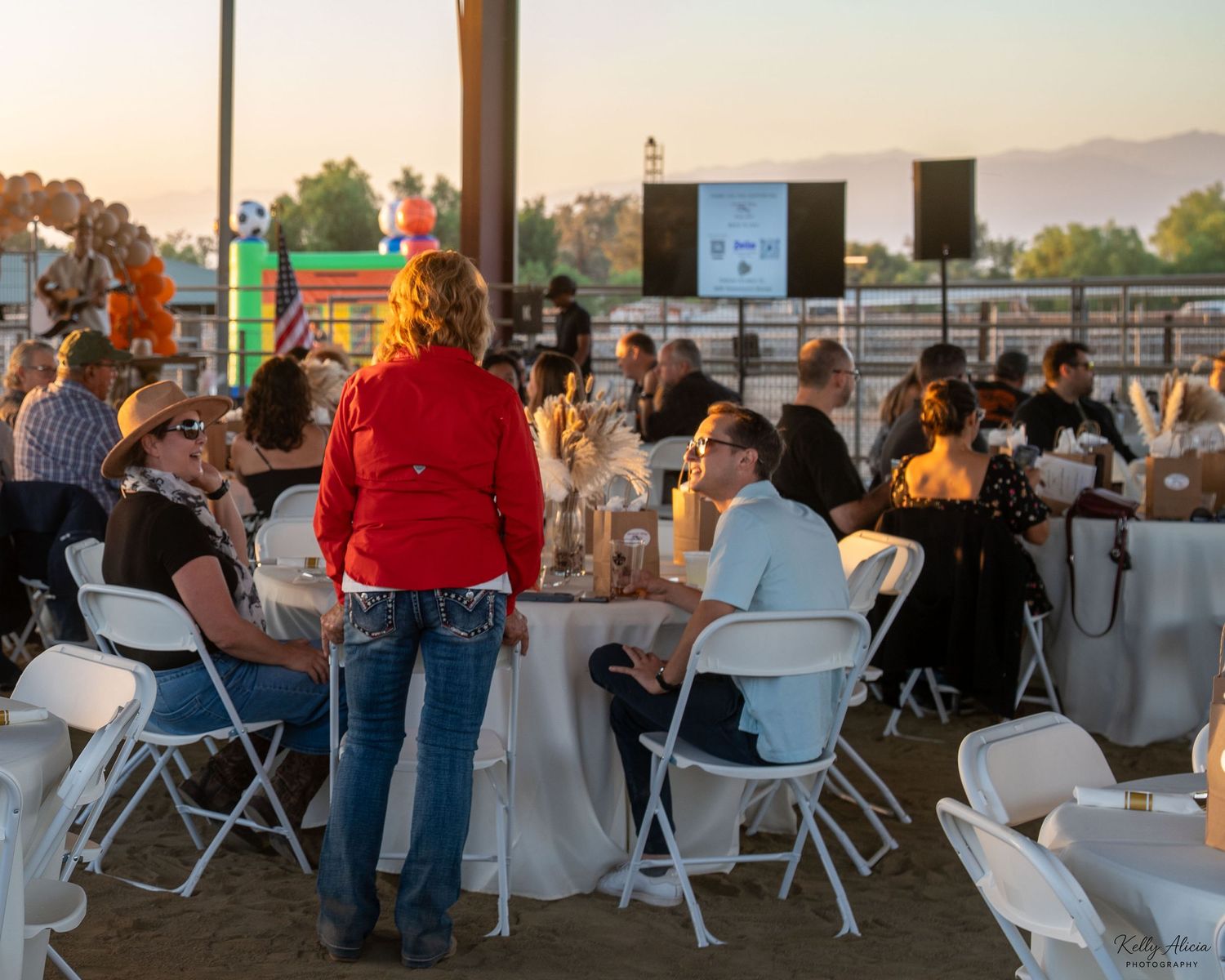 People gather at an outdoor event venue with folding chairs and tables during sunset against a mountain backdrop.