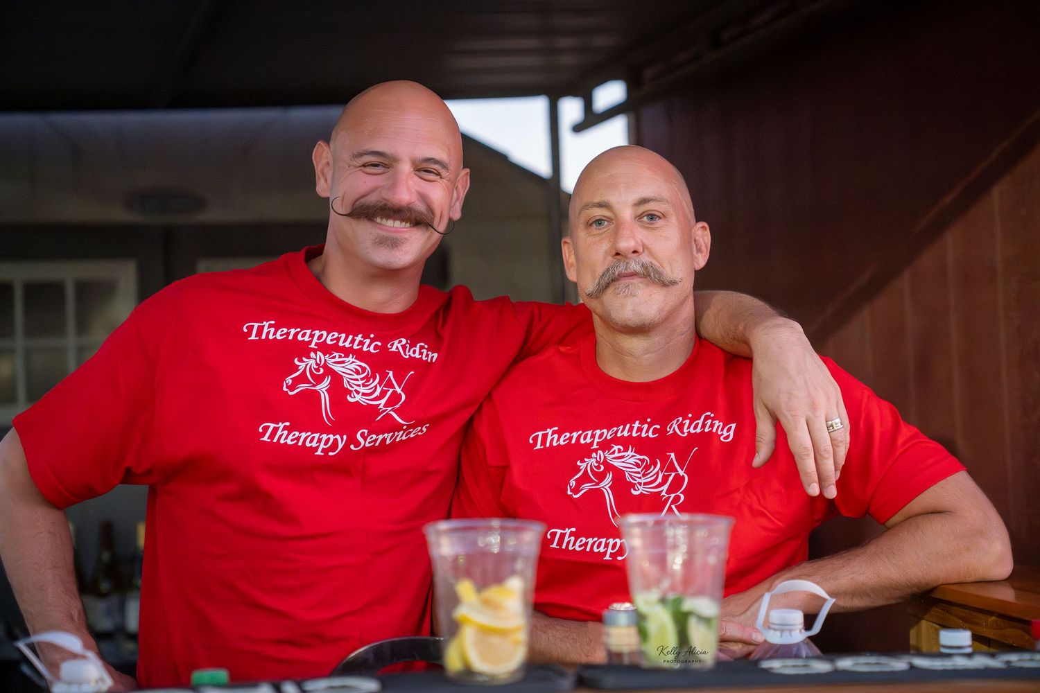 A group of people in matching red t-shirts gather around a table with drinks and snacks at a casual event.