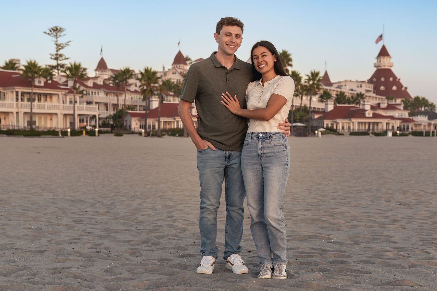 A couple poses together on a beach with historic Victorian buildings visible in the background.