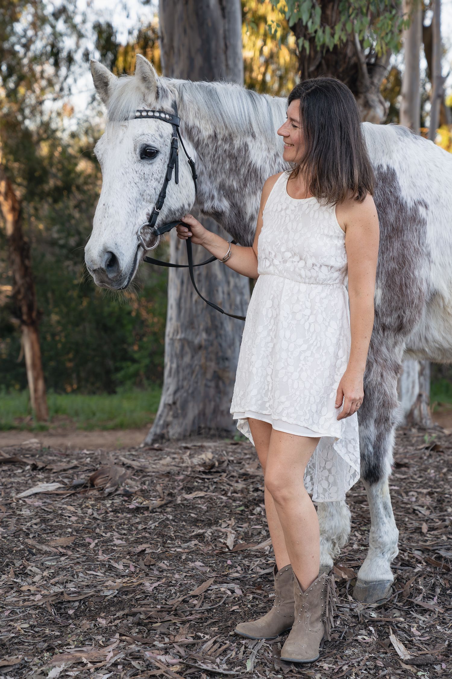 Woman in white dress standing next to a beautiful grey horse in a eucalyptus grove.