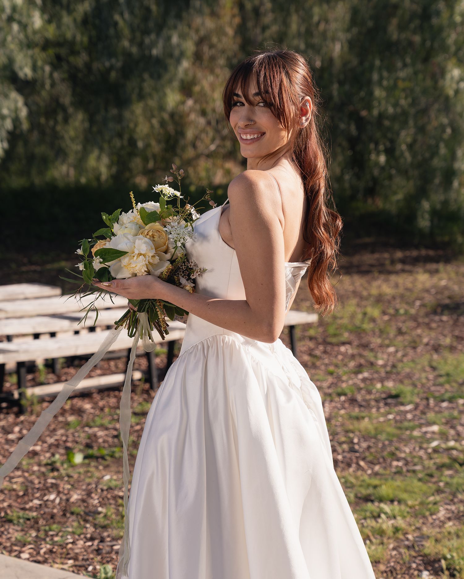 Smiling bride in white strapless gown holding white floral bouquet outdoors with trees in background.