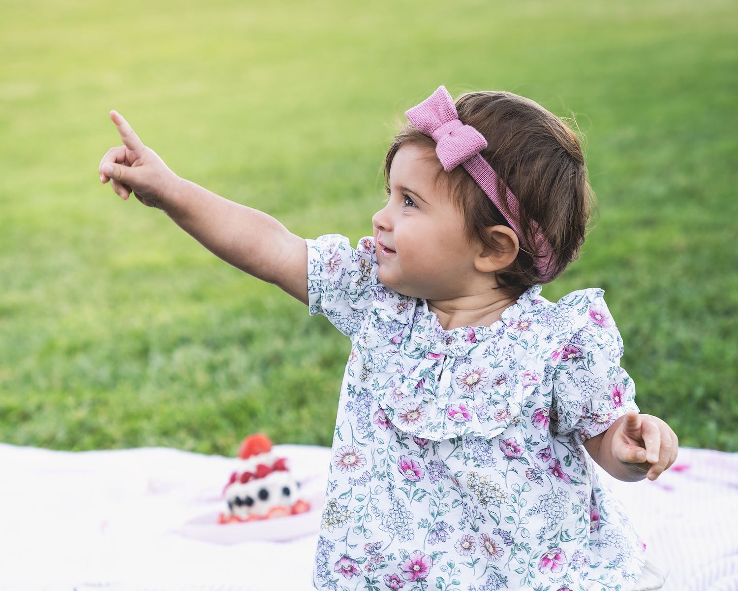 A small child in floral outfit points upward while sitting on picnic blanket outdoors.
