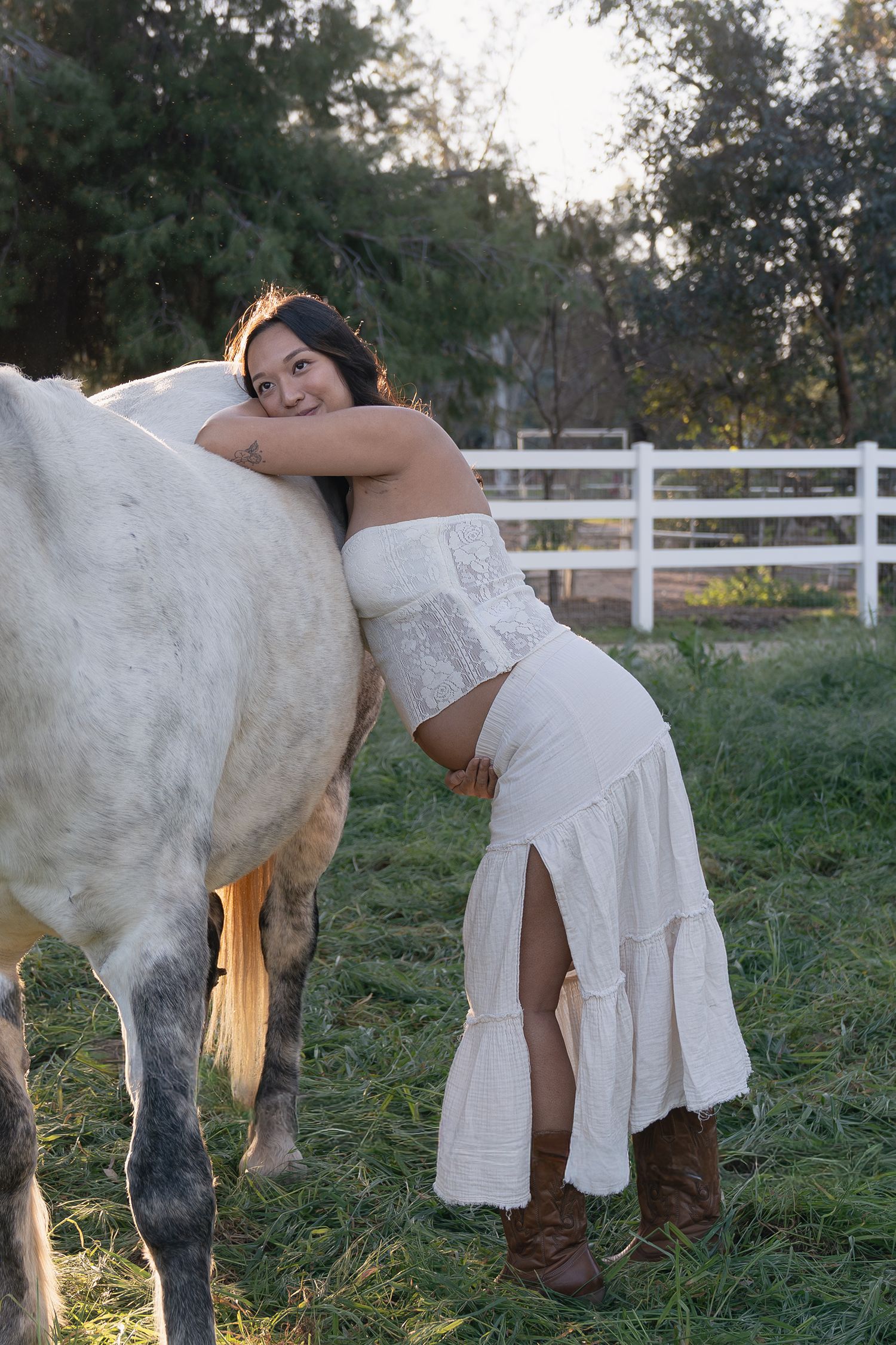 A person in a white dress stands beside a white horse in a pastoral field with white fencing at sunset.