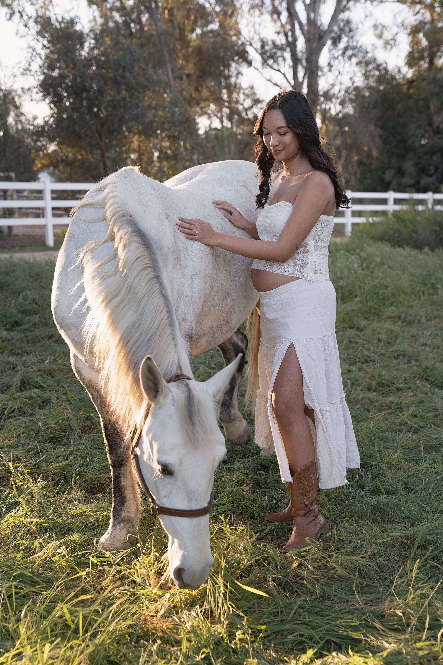 A person in a flowing white dress stands beside a white horse grazing in a sunlit meadow at golden hour.