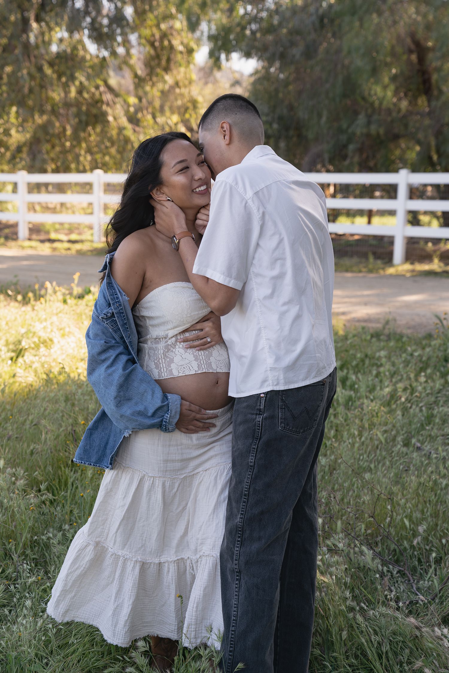 A couple shares an intimate moment in a field with a white fence and sunlit trees in the background.