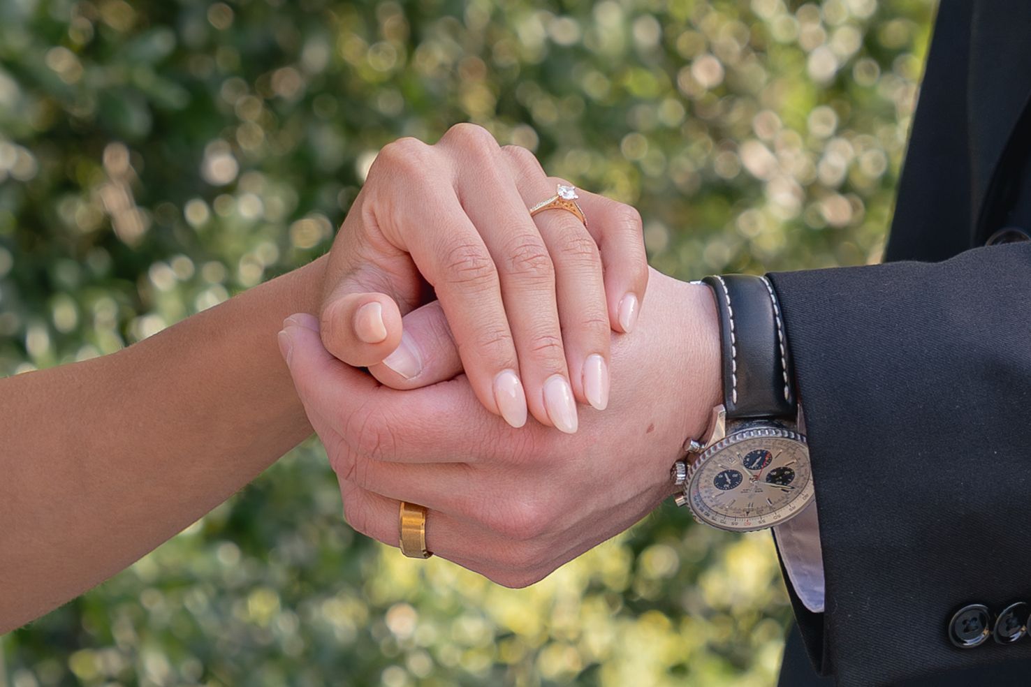 Couple holding hands showing wedding rings and a luxury watch, with green bokeh background.