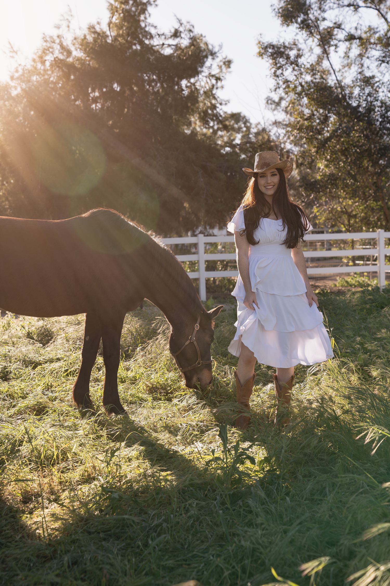 A person in a white dress stands next to a dark horse in a sunlit field with white fencing in the background.