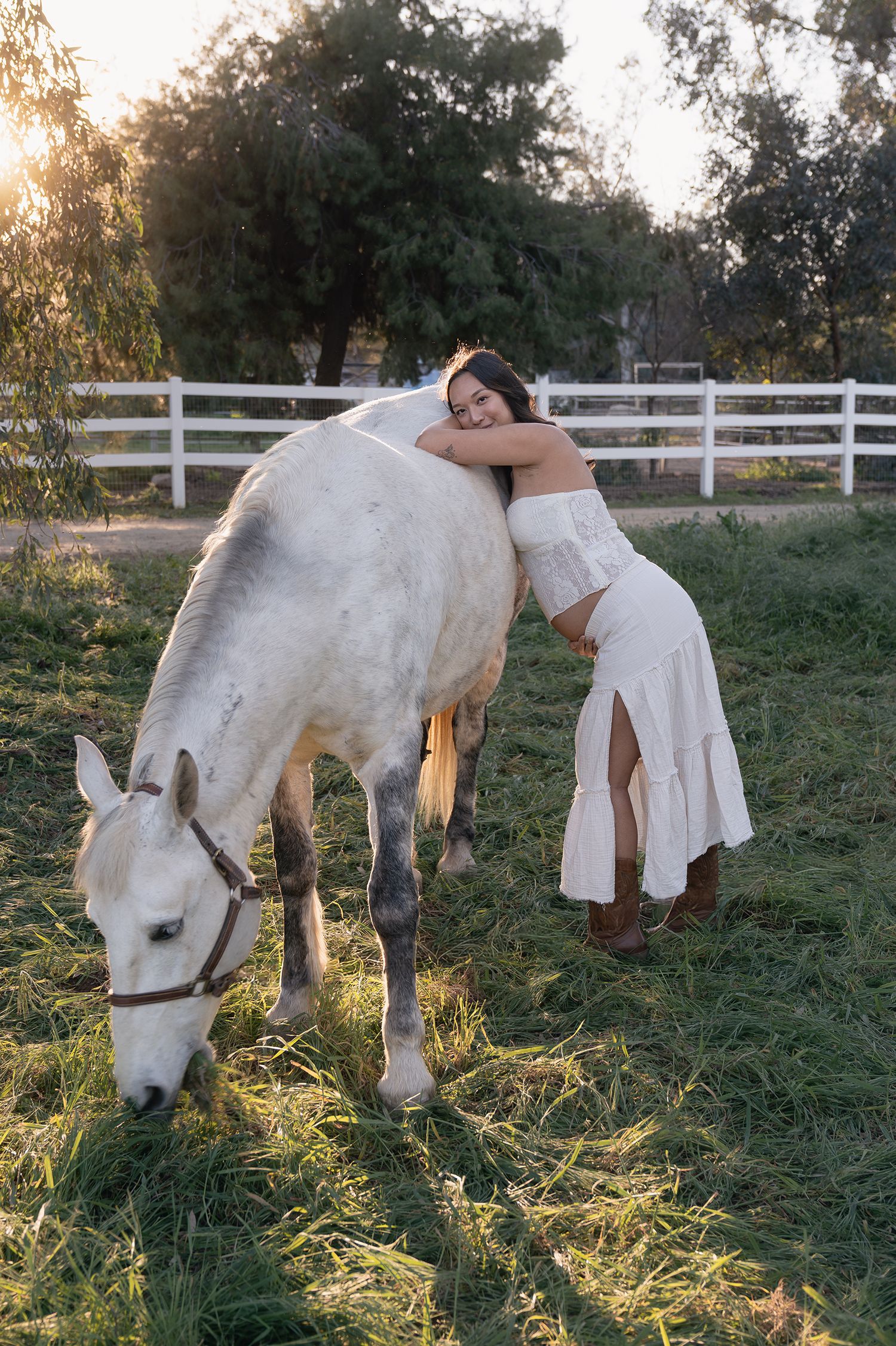 A white horse grazes in a sunlit pasture with white fencing while someone in a flowing white dress leans on its back.