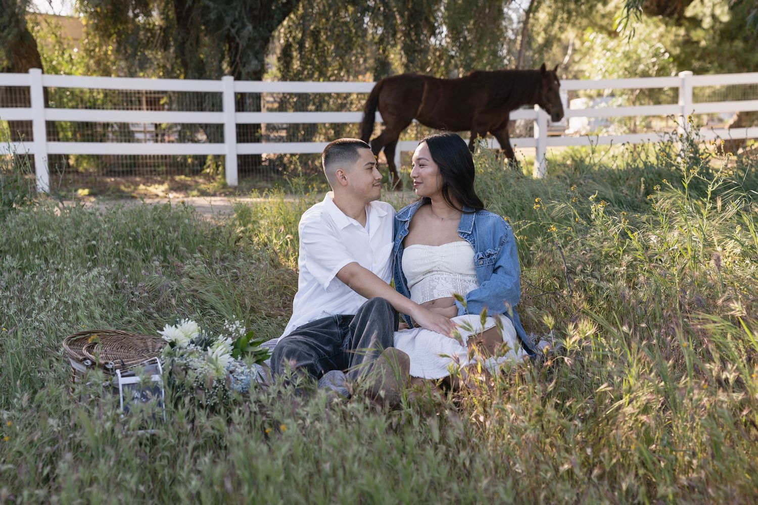 A couple shares a romantic moment in a rustic outdoor setting with white fencing and natural foliage in the background.