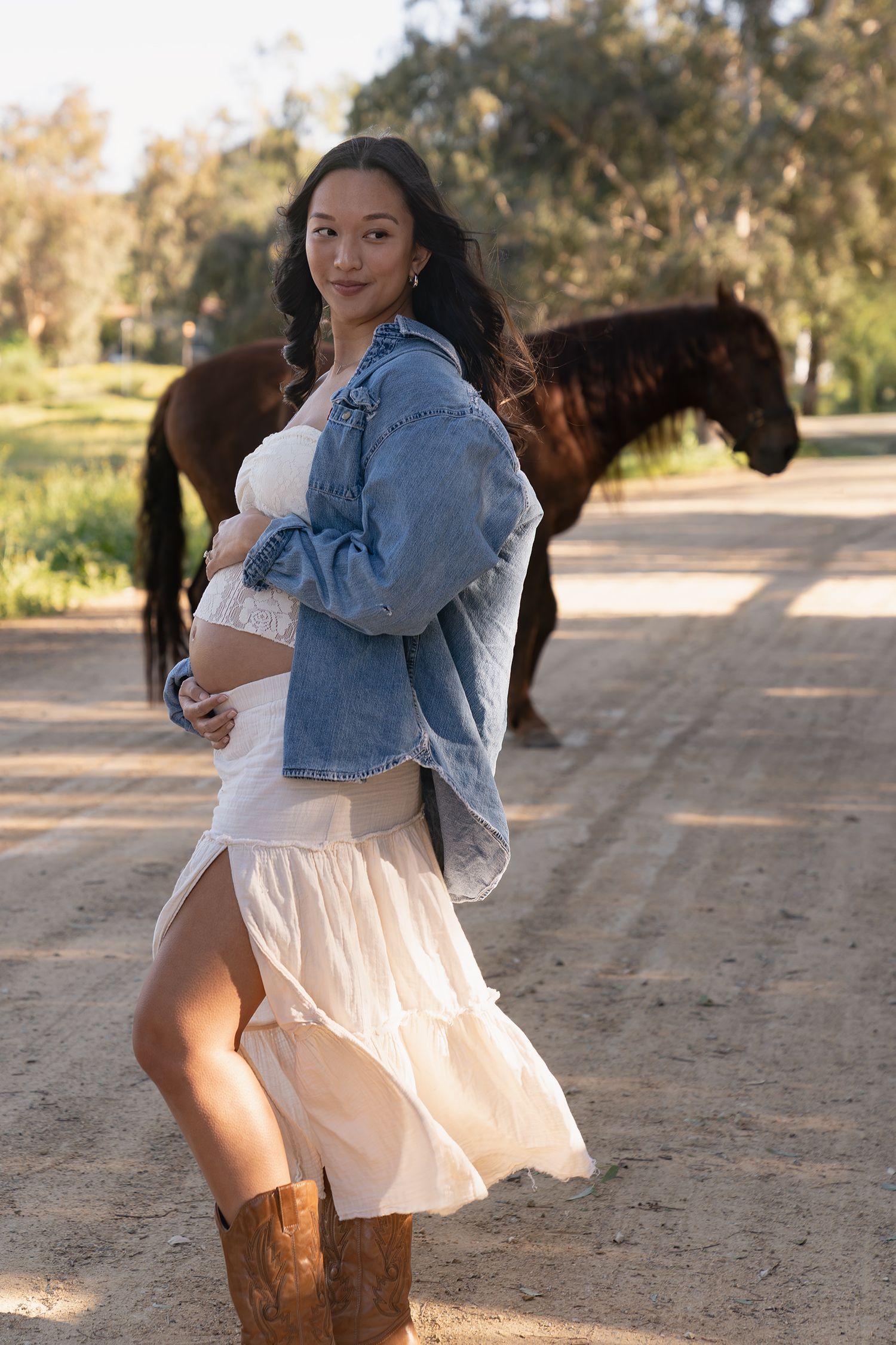 A person in a white dress and denim jacket spins playfully in front of a horse on a dirt path during golden hour.