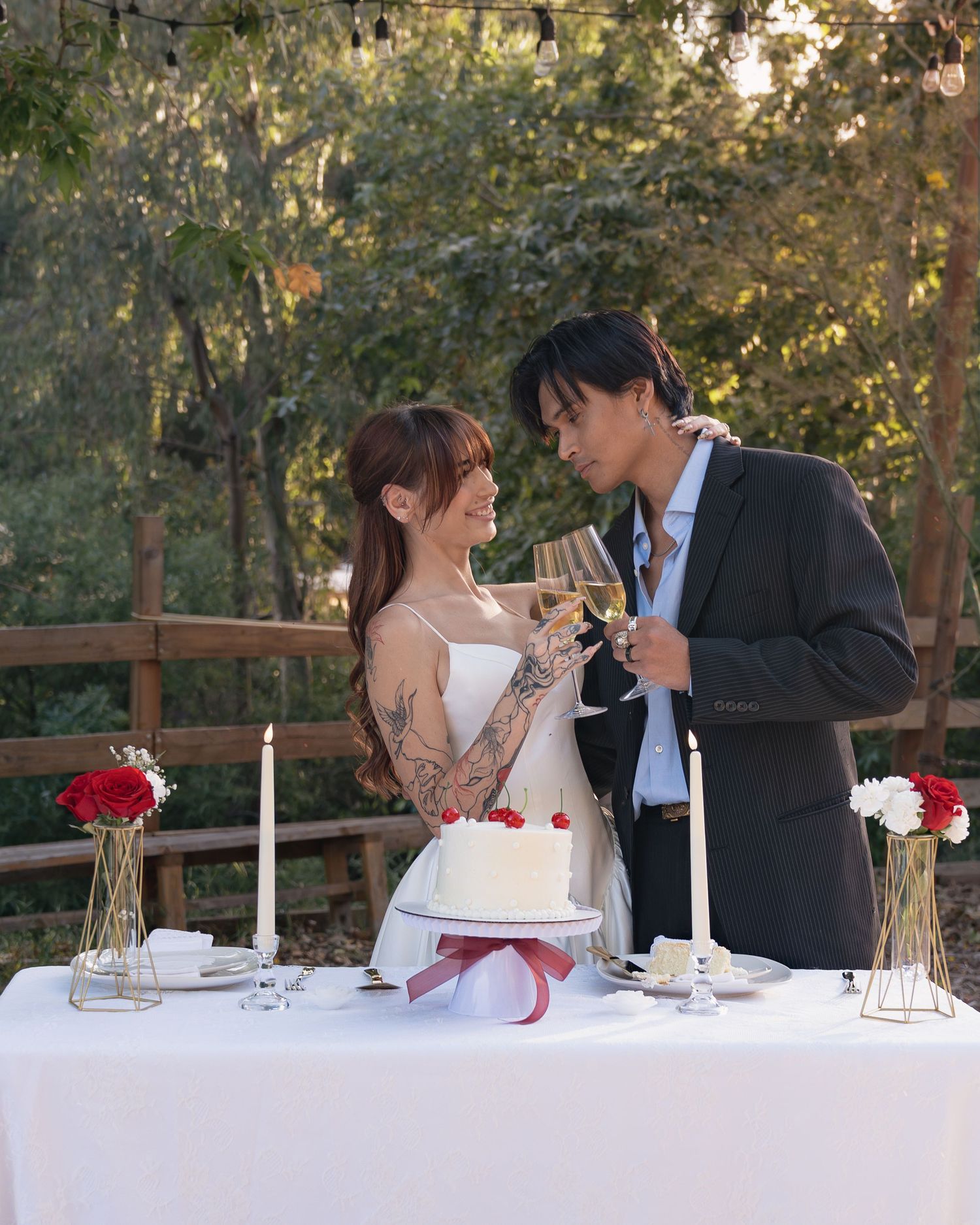 Couple toasting with champagne at outdoor wedding table with white cake, candles, and red roses.