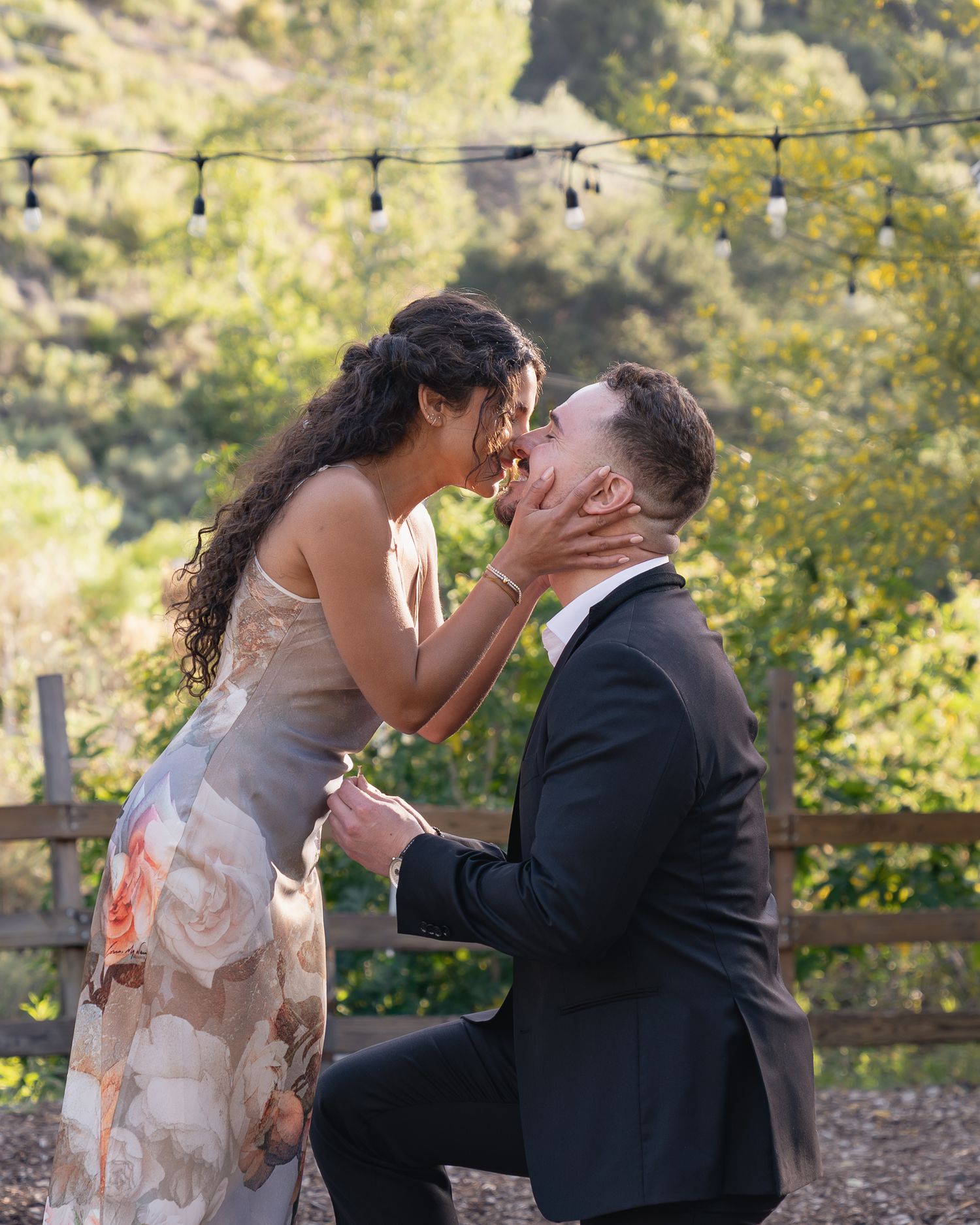 A man kneels on one knee proposing to a woman in a floral dress outdoors with string lights and greenery.