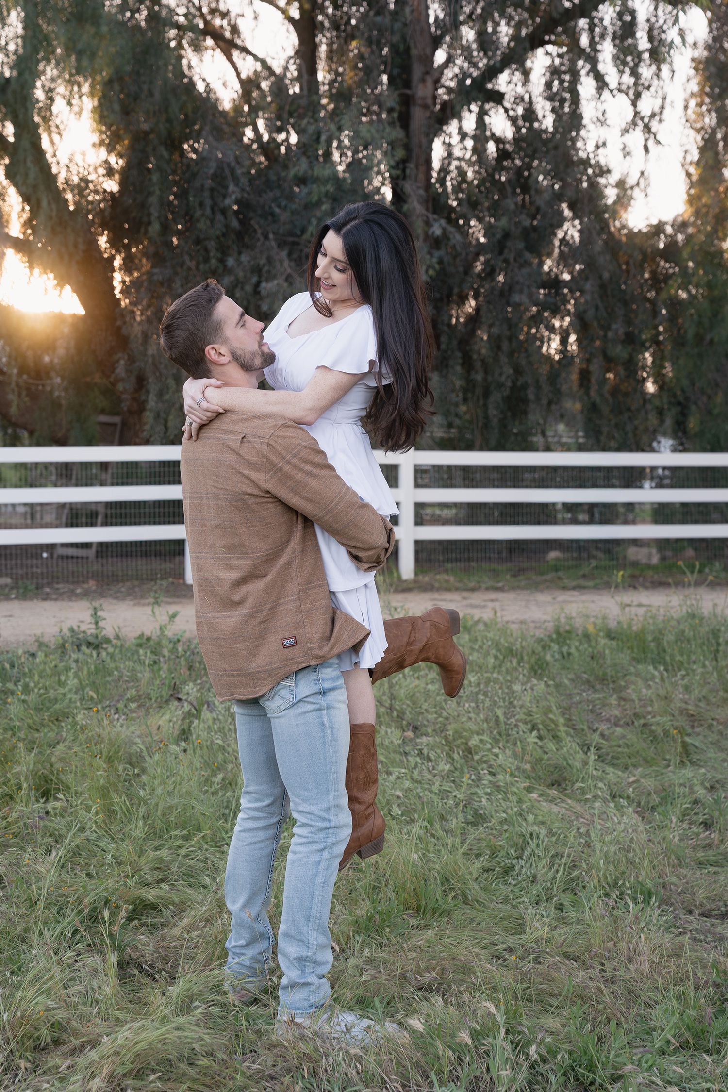 A couple shares a romantic embrace in a field at sunset with white fencing visible in the background.