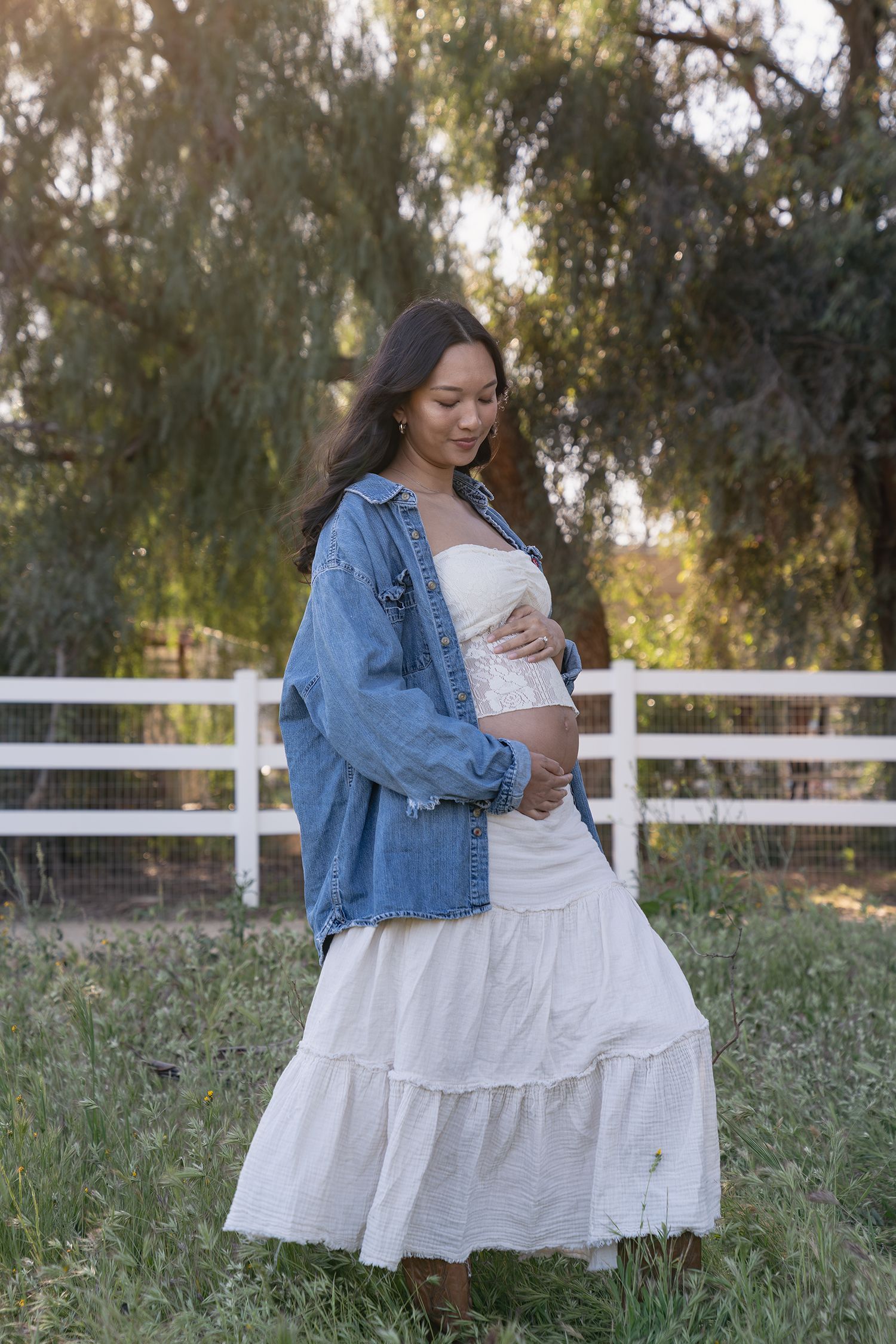 Woman in white flowy dress and denim jacket poses in outdoor field with white fence at sunset.