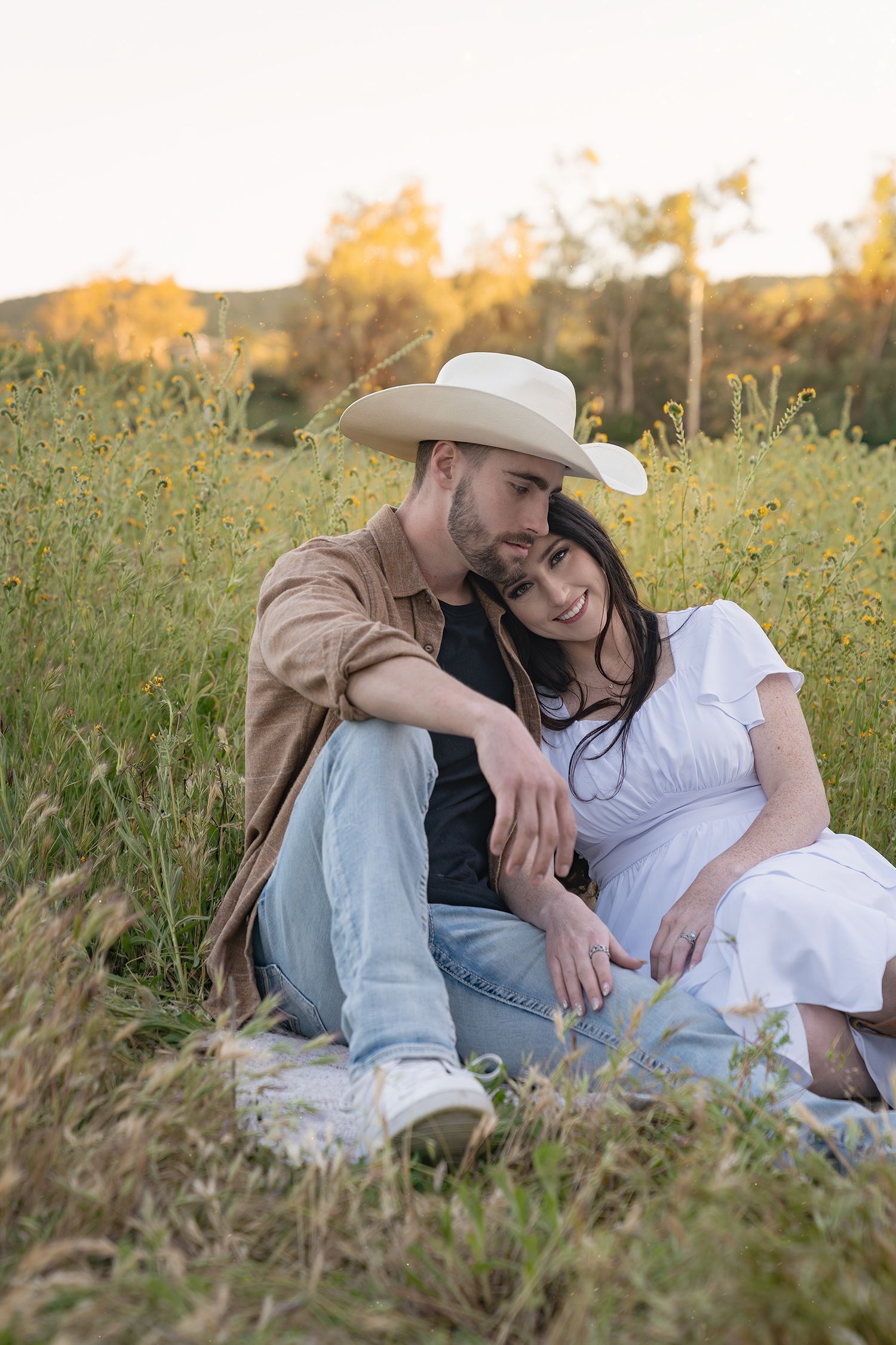 A couple sits together in a wildflower meadow at sunset, with the person in a cowboy hat and brown shirt embracing their partner.