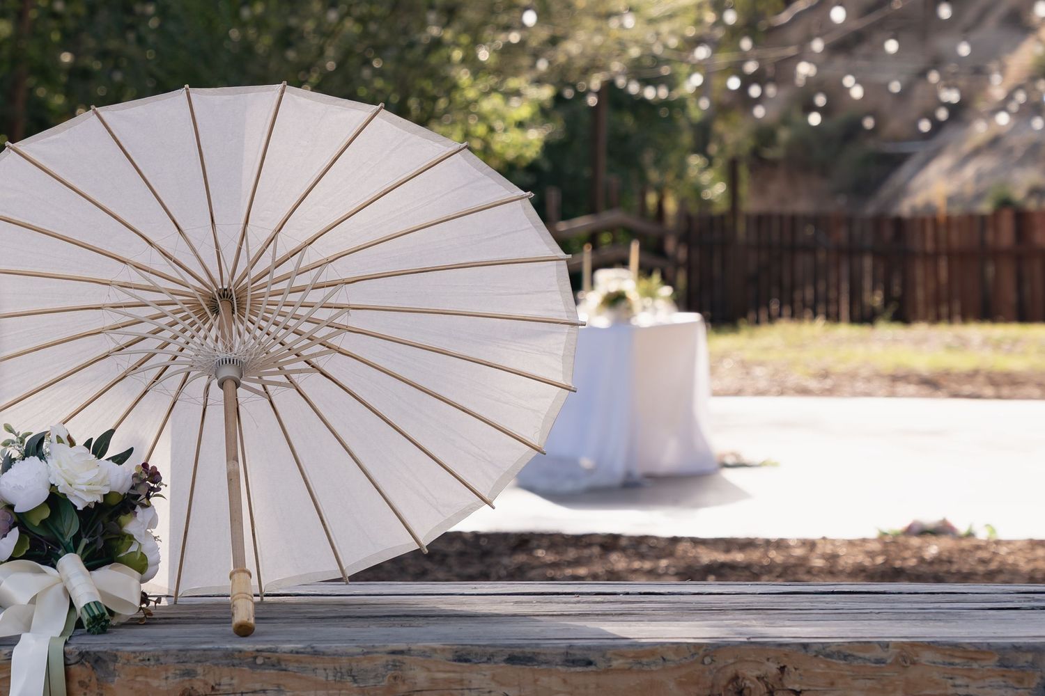White paper parasol and floral bouquet on wooden bench at outdoor wedding venue with string lights.