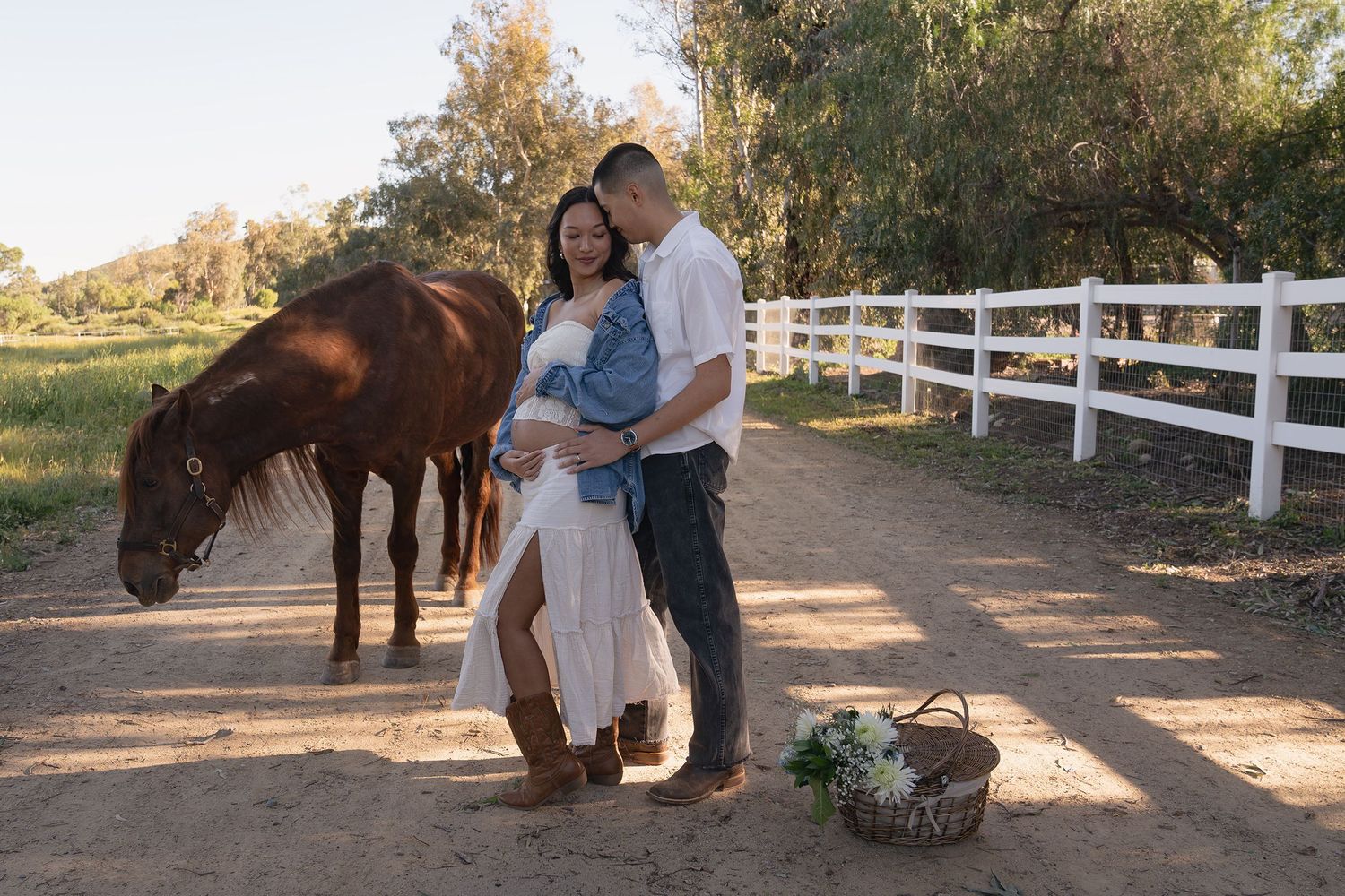 Couple poses with horses at sunset on a ranch with white fencing and autumn trees in the background.