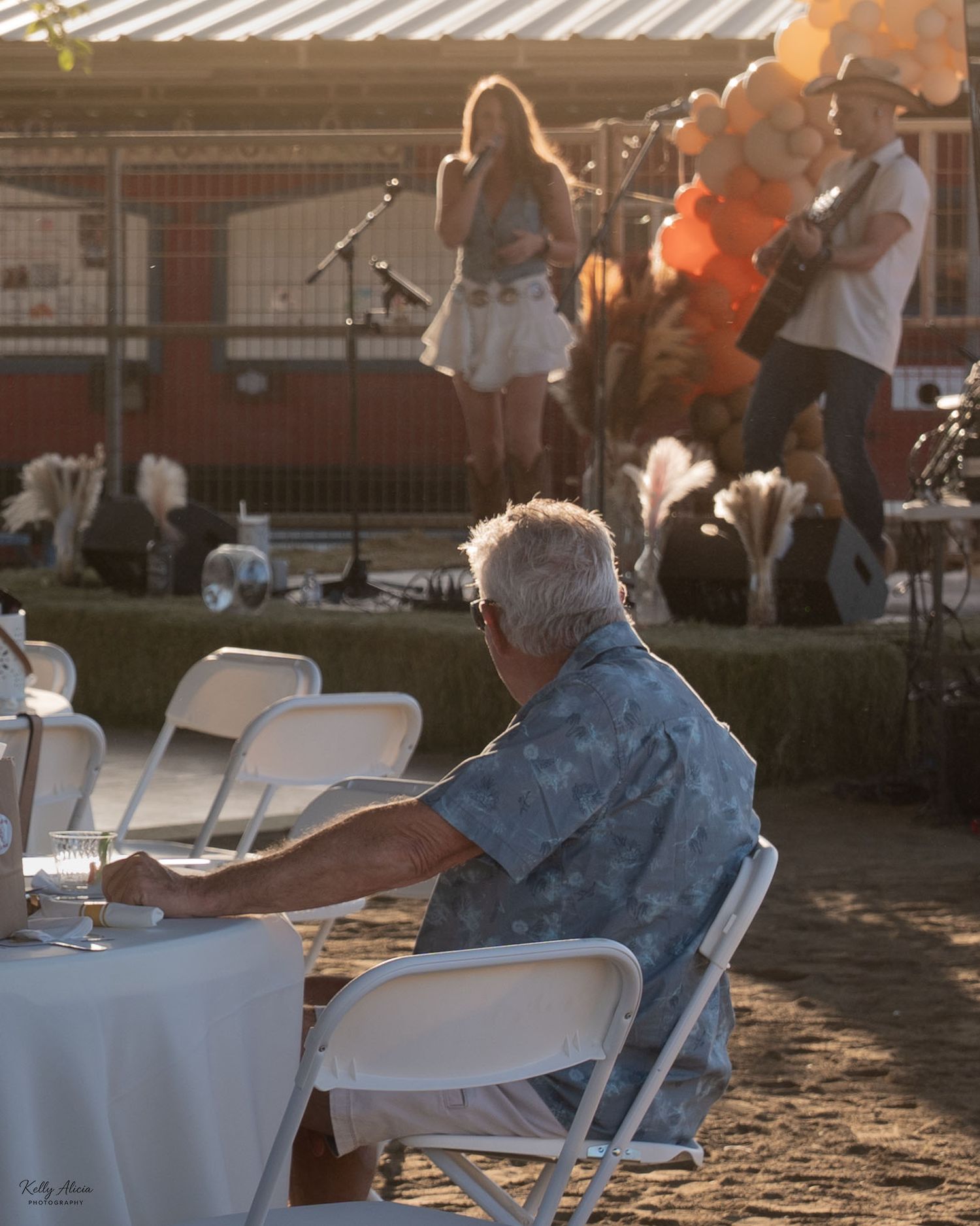 People sit in white chairs during a golden sunset as light streams through a slatted pergola structure.
