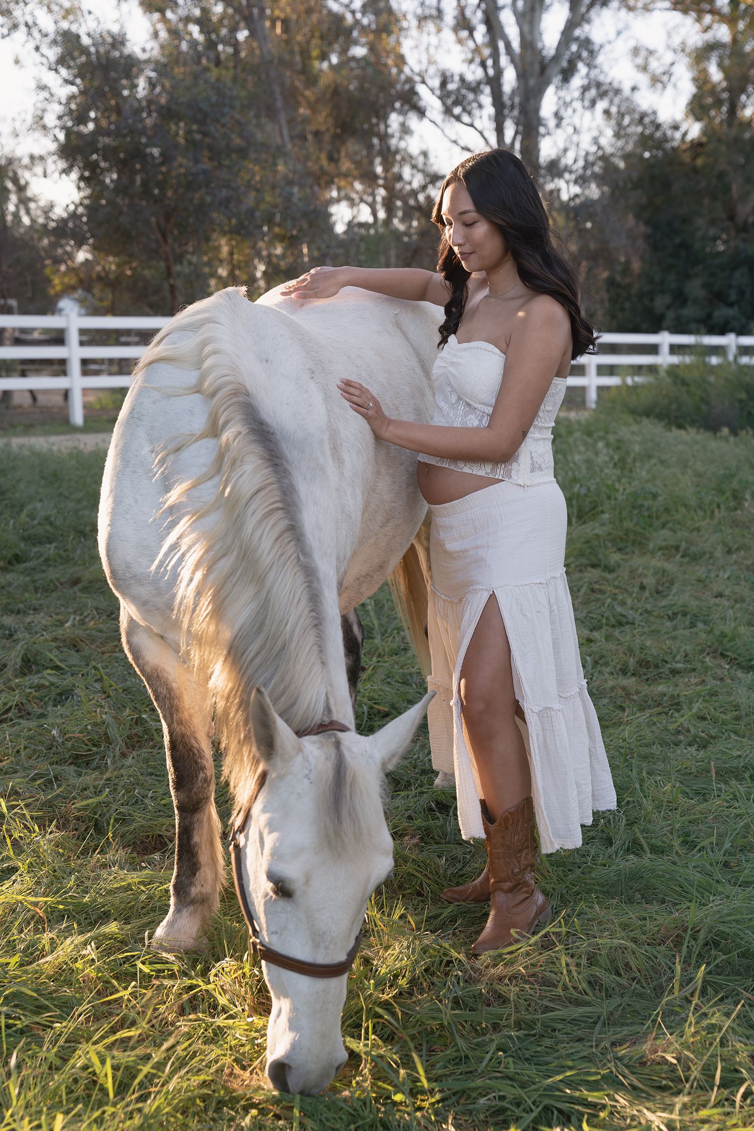 A white horse grazes in a sunlit field while someone in a flowing white dress gently pets its mane.