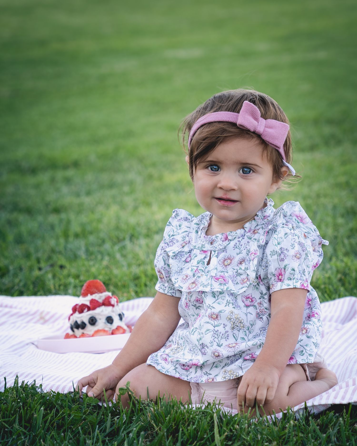 A toddler in floral dress and pink bow sitting on blanket with dessert in background.