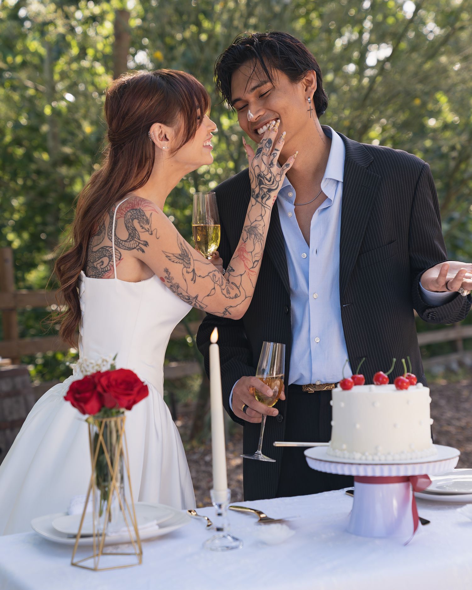 Tattooed bride in white dress and groom in navy suit share a romantic moment beside wedding cake and red roses.