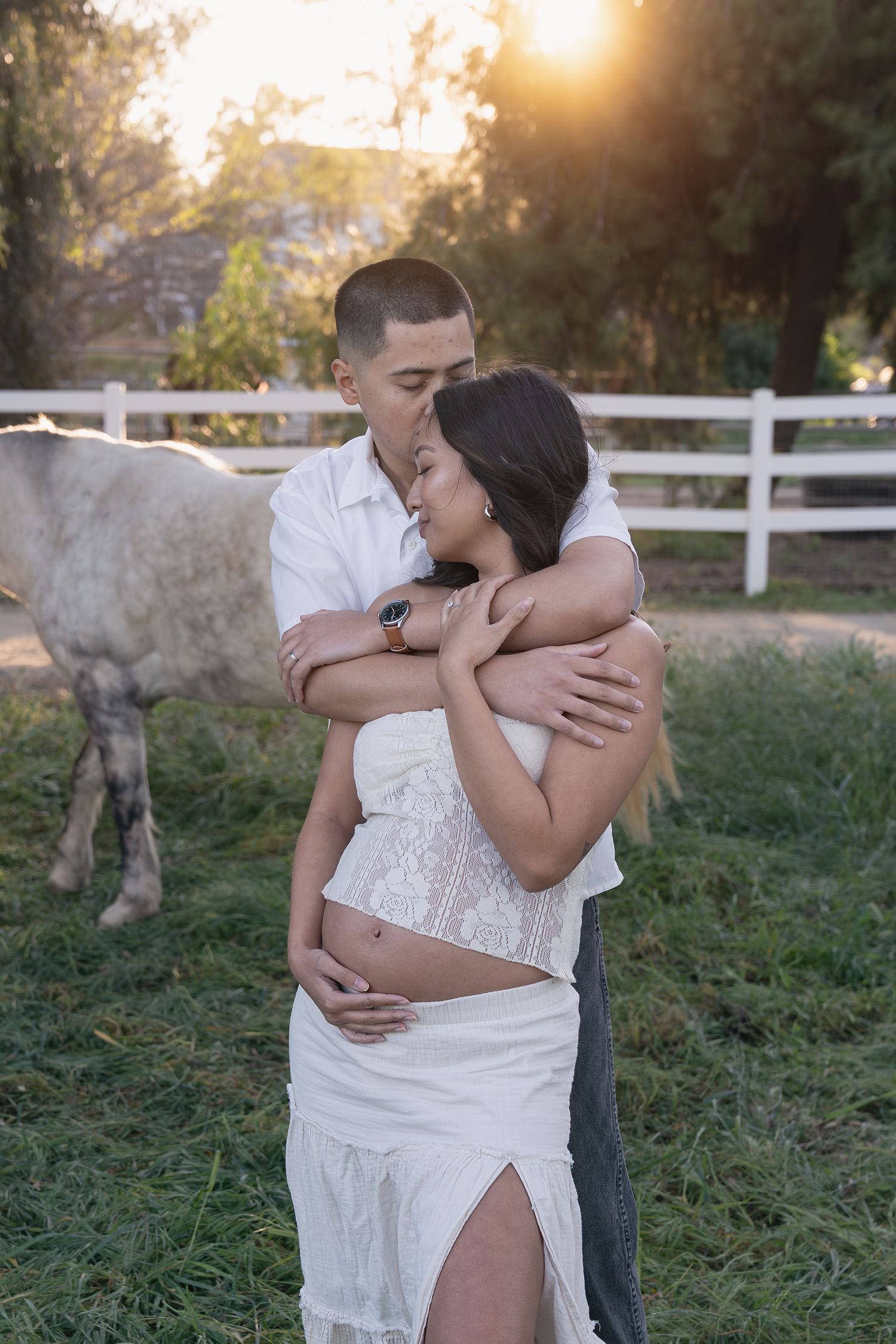 A romantic couple embraces in a sunlit field with a white fence and horse in the background at sunset.