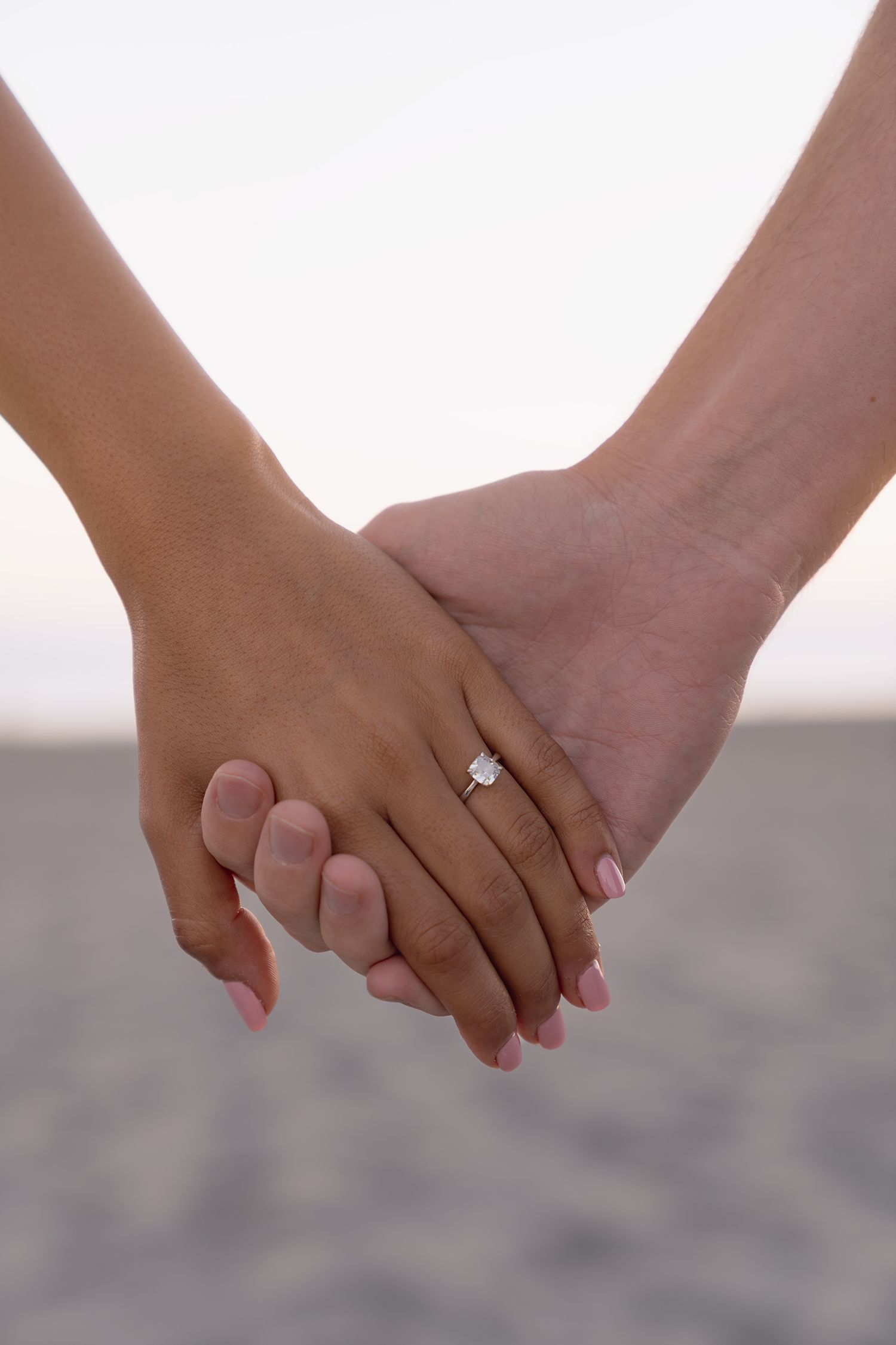 Two hands hold each other tenderly with an engagement ring visible against a soft blurred background.