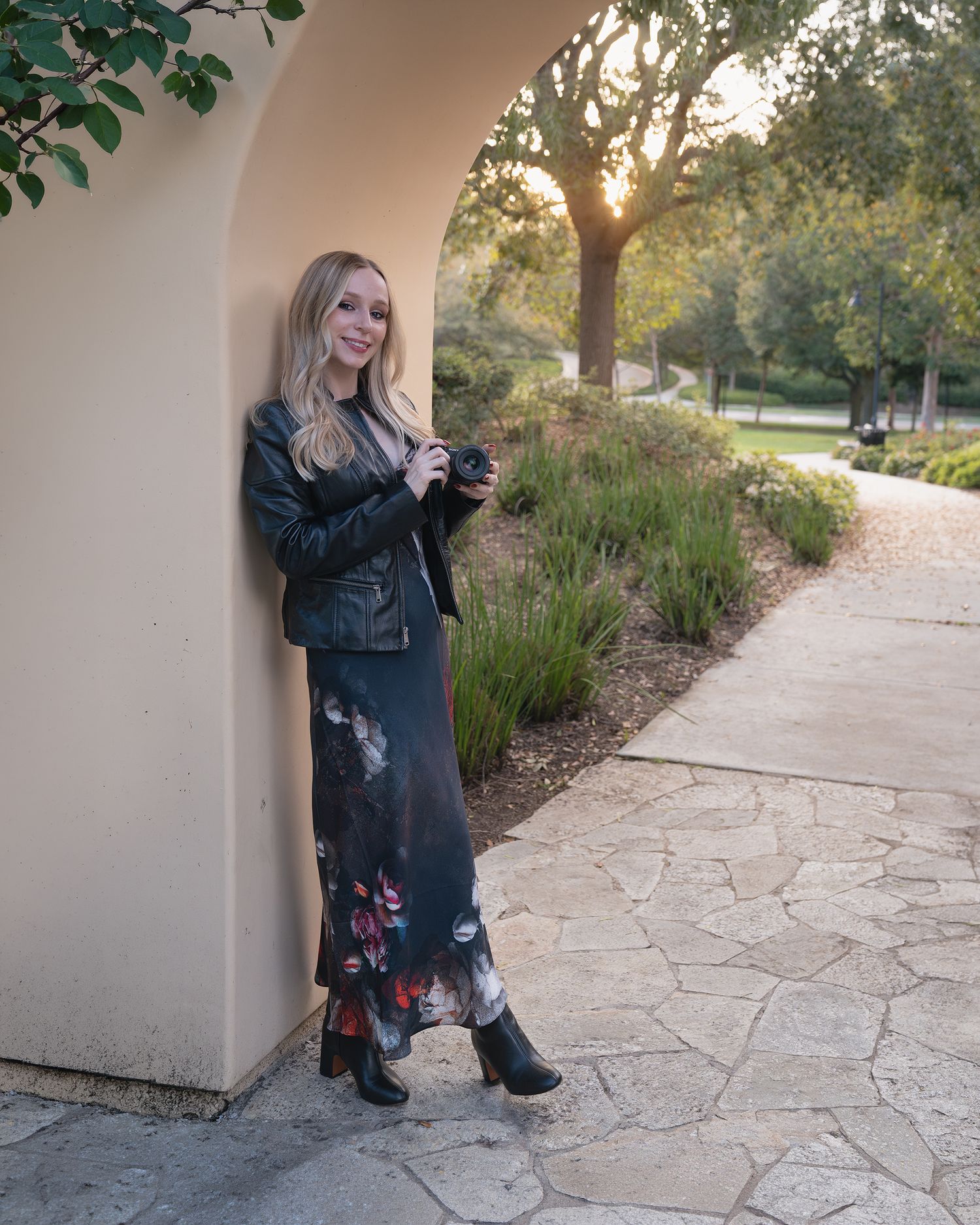 A person in a leather jacket and floral maxi dress poses against a white wall on a stone path at sunset.