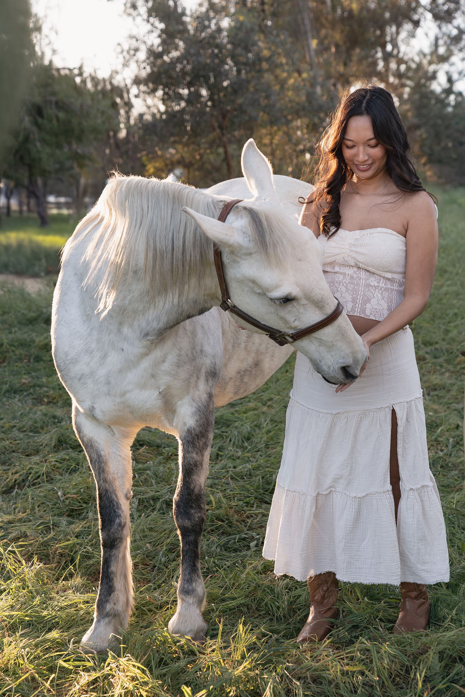 A white pony and someone in a white dress interact in a sunlit grassy field during golden hour.