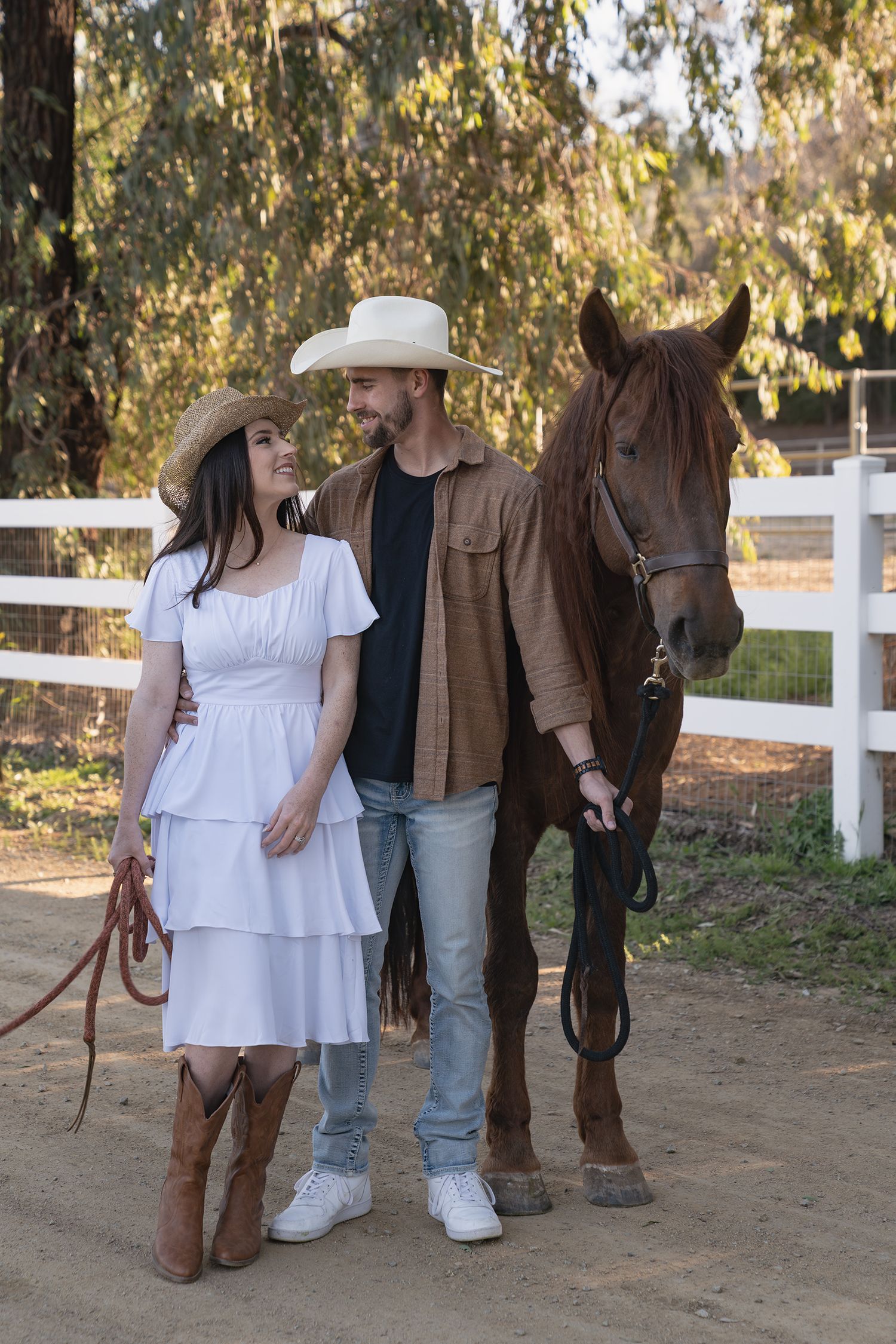 A couple stands with their brown horse against a white fence while wearing casual western-inspired outfits.