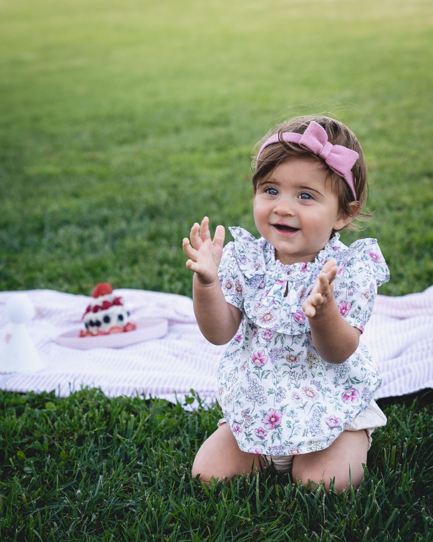 Happy child in floral dress and pink bow sitting on grass during outdoor photoshoot.