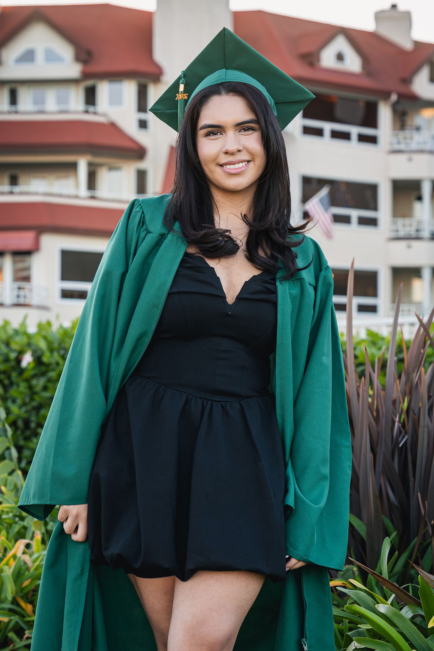 A person wearing a green graduation gown and black dress poses outdoors in front of residential buildings.