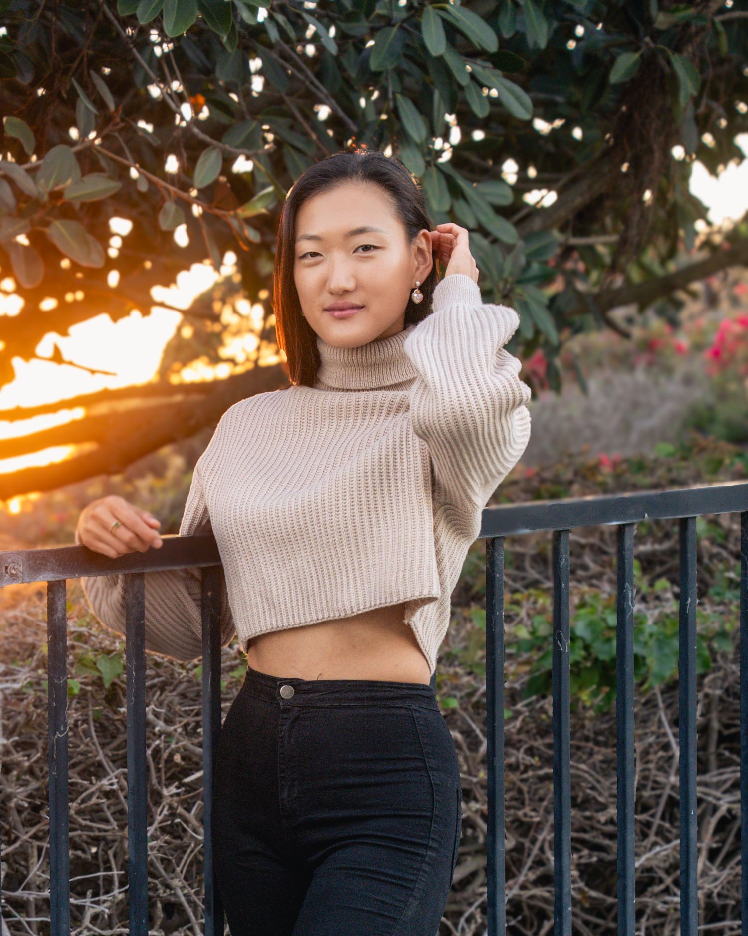 Person in beige sweater and dark pants posing against sunset with foliage in background.