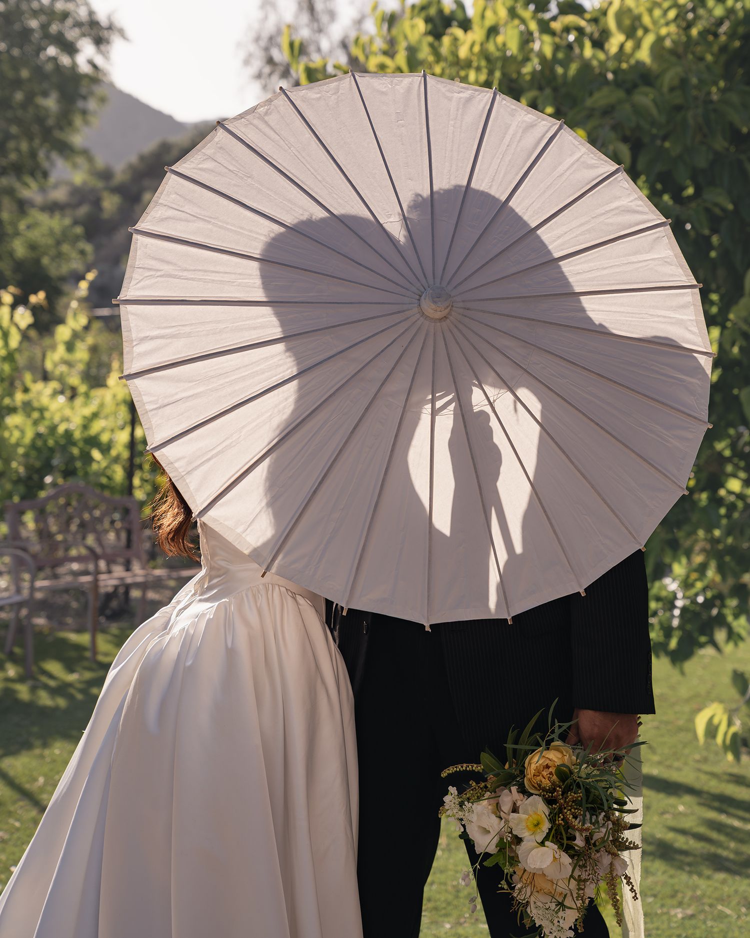A wedding couple kissing behind a white parasol umbrella, their silhouette forming a heart shape, outdoors in a garden.