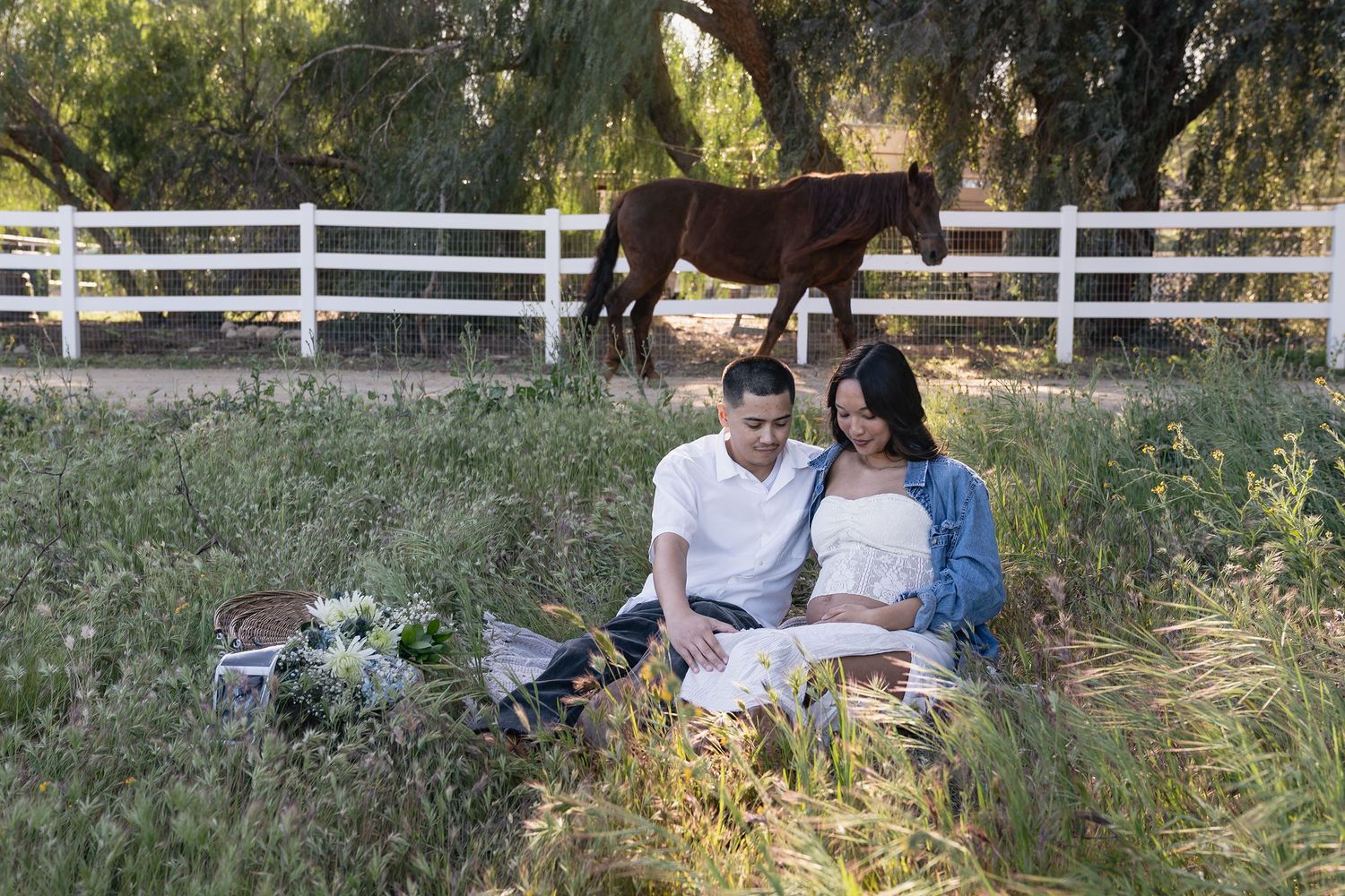 A couple sits together on a rustic wooden bench surrounded by tall grass and white fencing in a romantic outdoor setting.